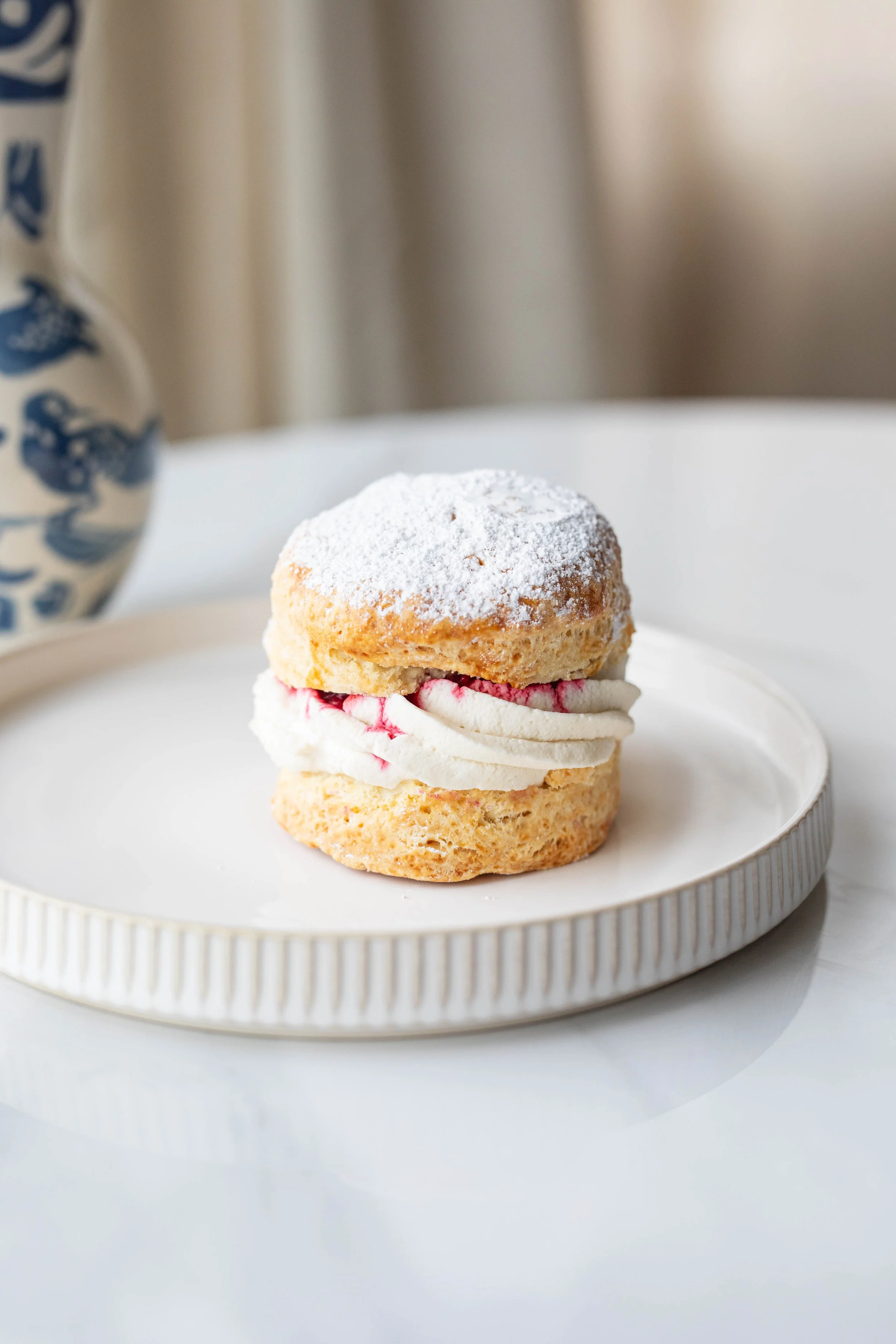 A close-up of a dessert with a choux pastry topped with powdered sugar, layered with cream and red fruit filling, on a white ridged plate.