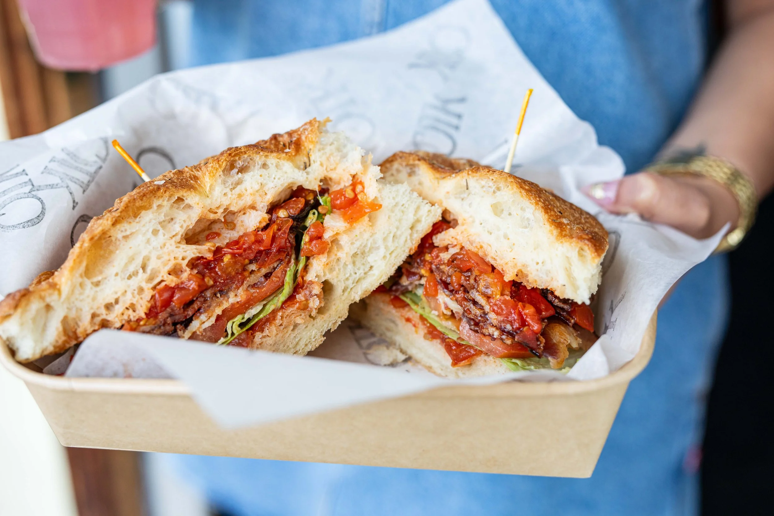 A sandwich sandwich with bread, lettuce, tomato, bacon, and possibly other ingredients, cut in half and served in a paper-lined container.