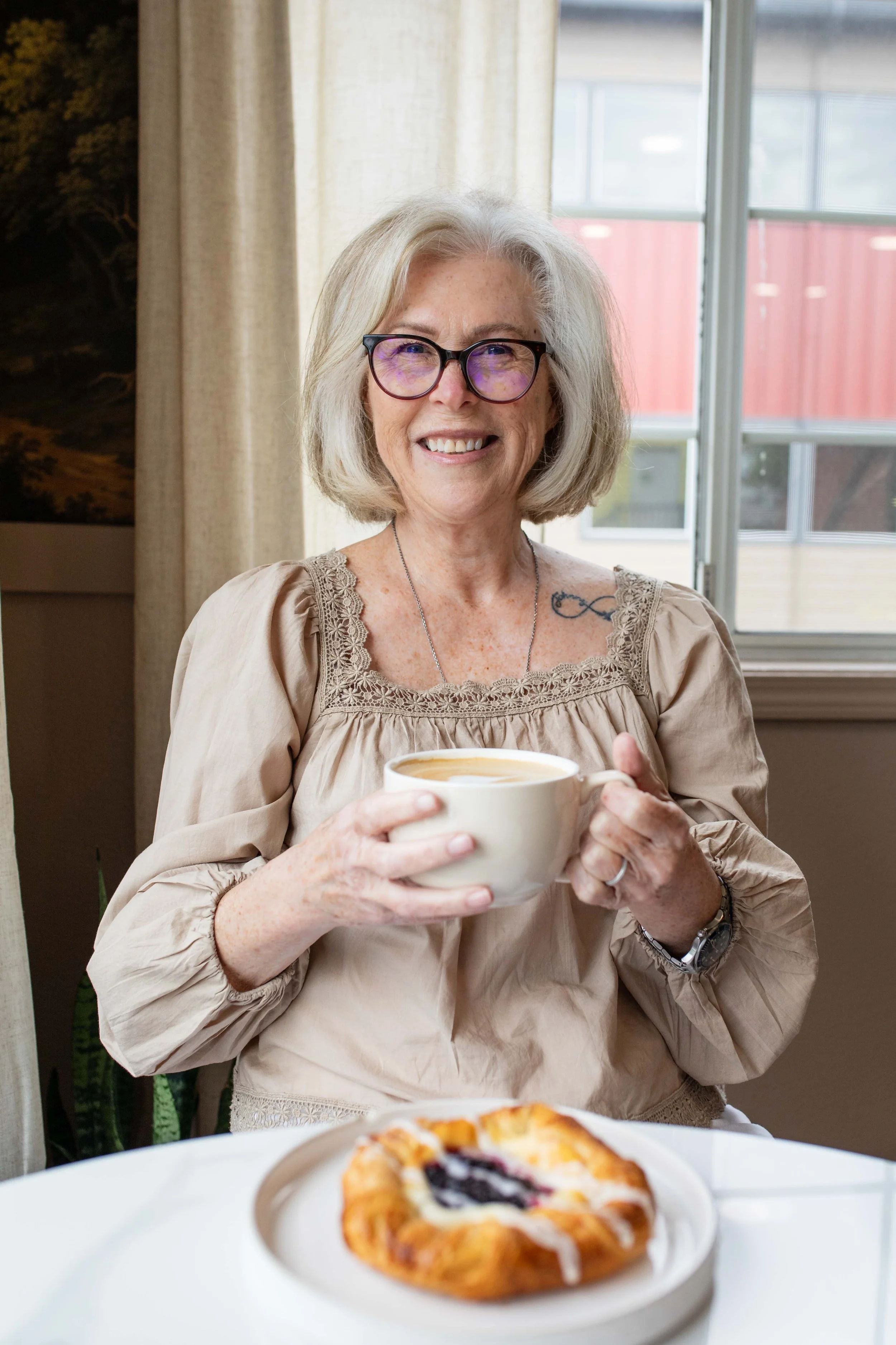 An woman with glasses and a beige blouse holds a cup of coffee, smiling at the camera, seated indoors near a window with curtains, with a slice of pie on the table in front of her.