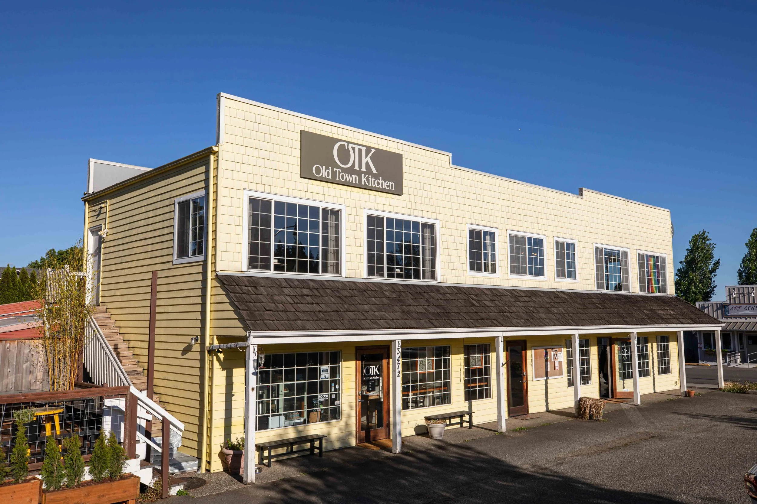 The exterior of Old Town Kitchen. The building is yellow with large windows, and there are outdoor benches and potted plants outside. The sky is clear and blue.
