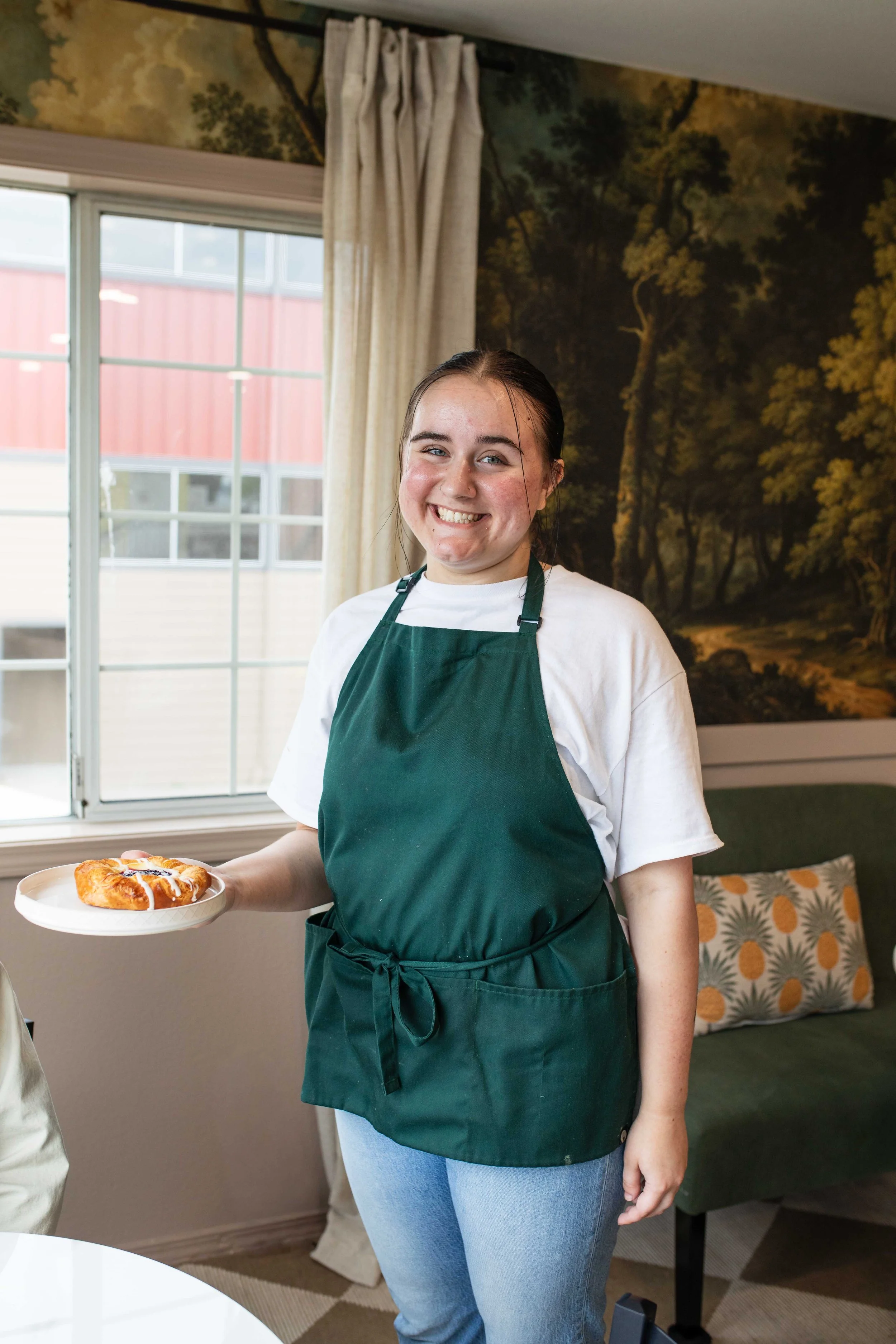 A young woman with dark hair tied back, smiling, wearing a white t-shirt and a green apron, holding a plate with a pastry. She is standing indoors near a window with cream-colored curtains, in a room with a mural of trees on the wall and a decorative pillow on a chair.