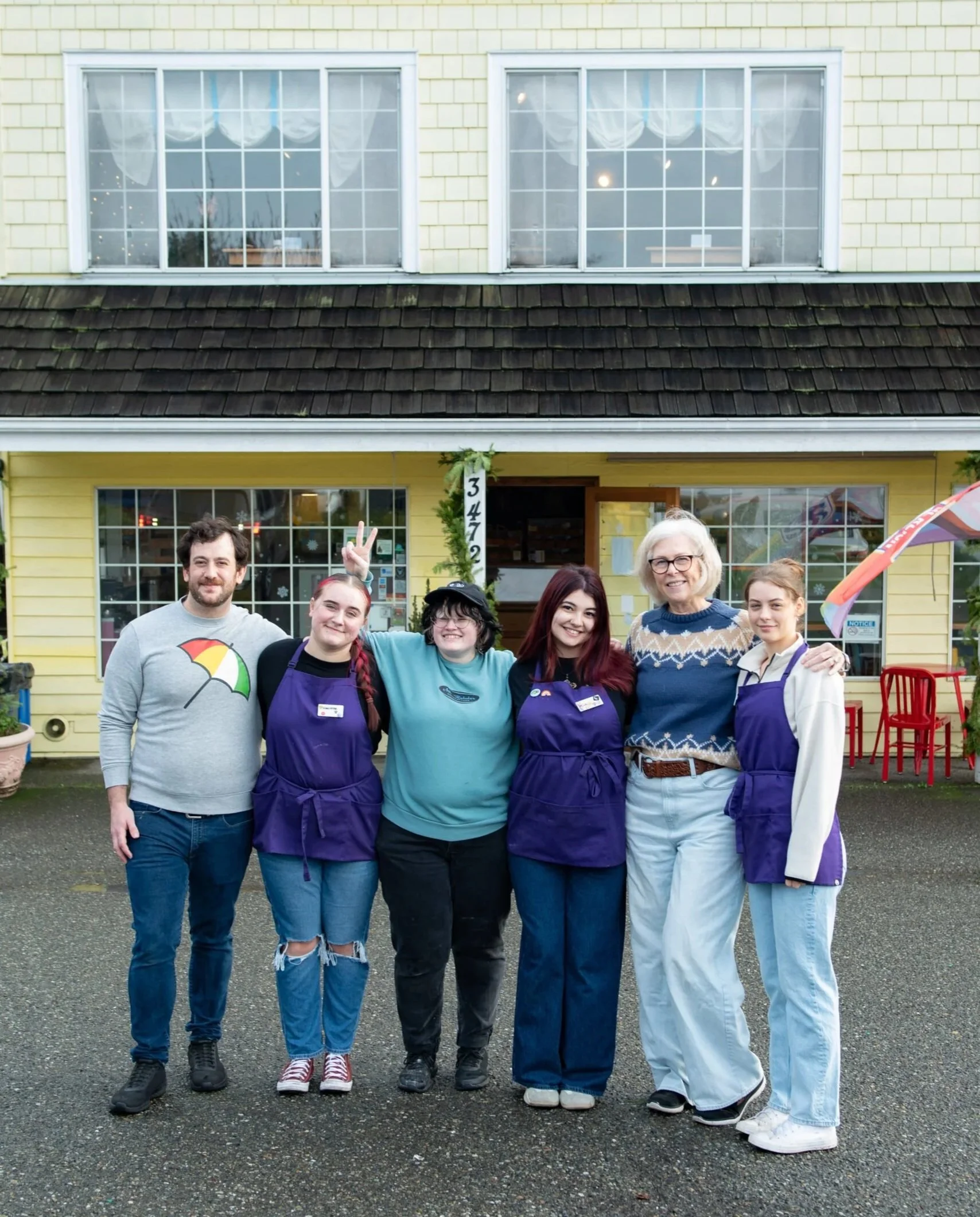 Group of six people standing in front of a yellow building with large windows, some wearing purple aprons, smiling and posing for the photo.