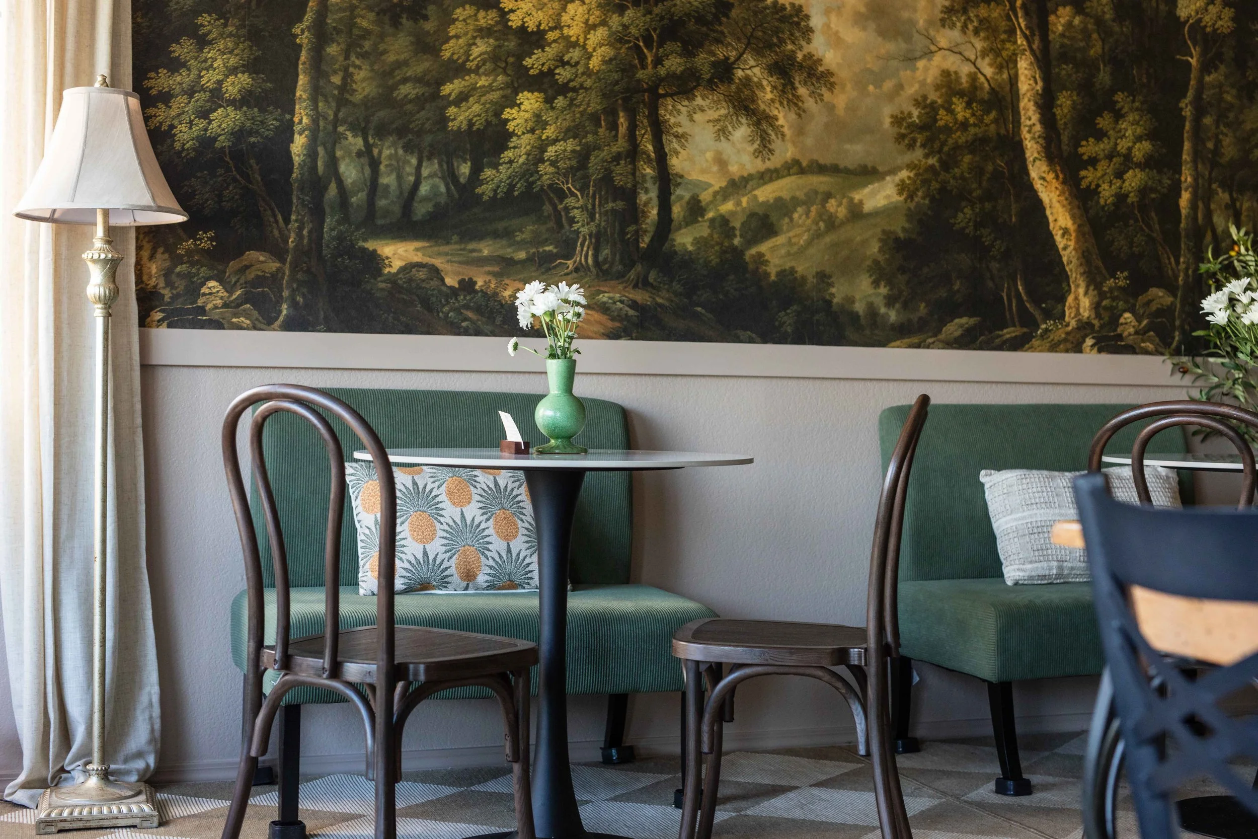 Interior of a dining area with green upholstered chairs around a table with a green vase holding white flowers, a tablecloth with pineapples, a patterned pillow, a standing lamp, and a large mural of a wooded landscape on the wall.