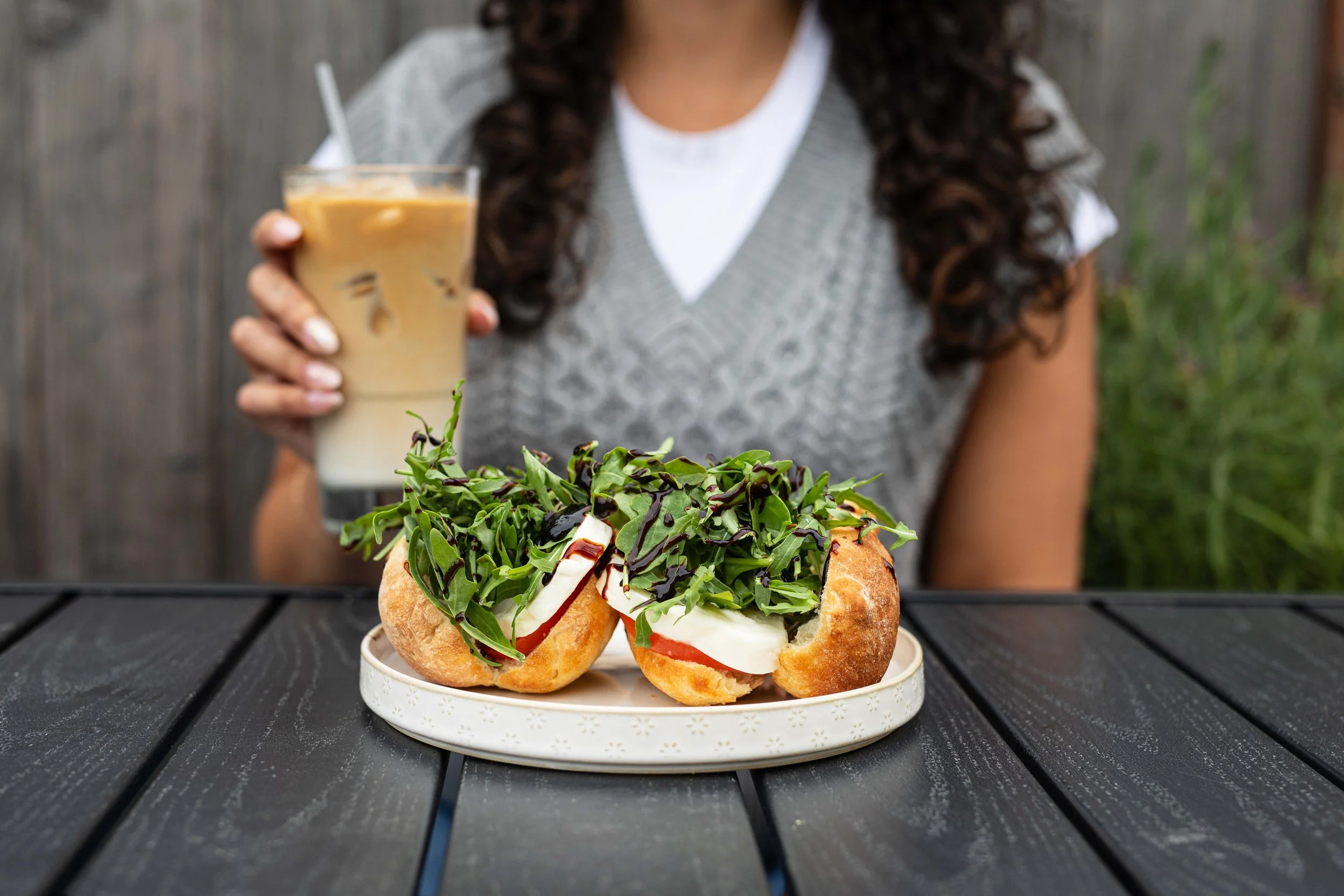 A sandwich with arugula, tomato, cheese, and drizzled with balsamic glaze, served on a bun, placed on a white plate on a black wooden table, with a person holding an iced coffee in the background.