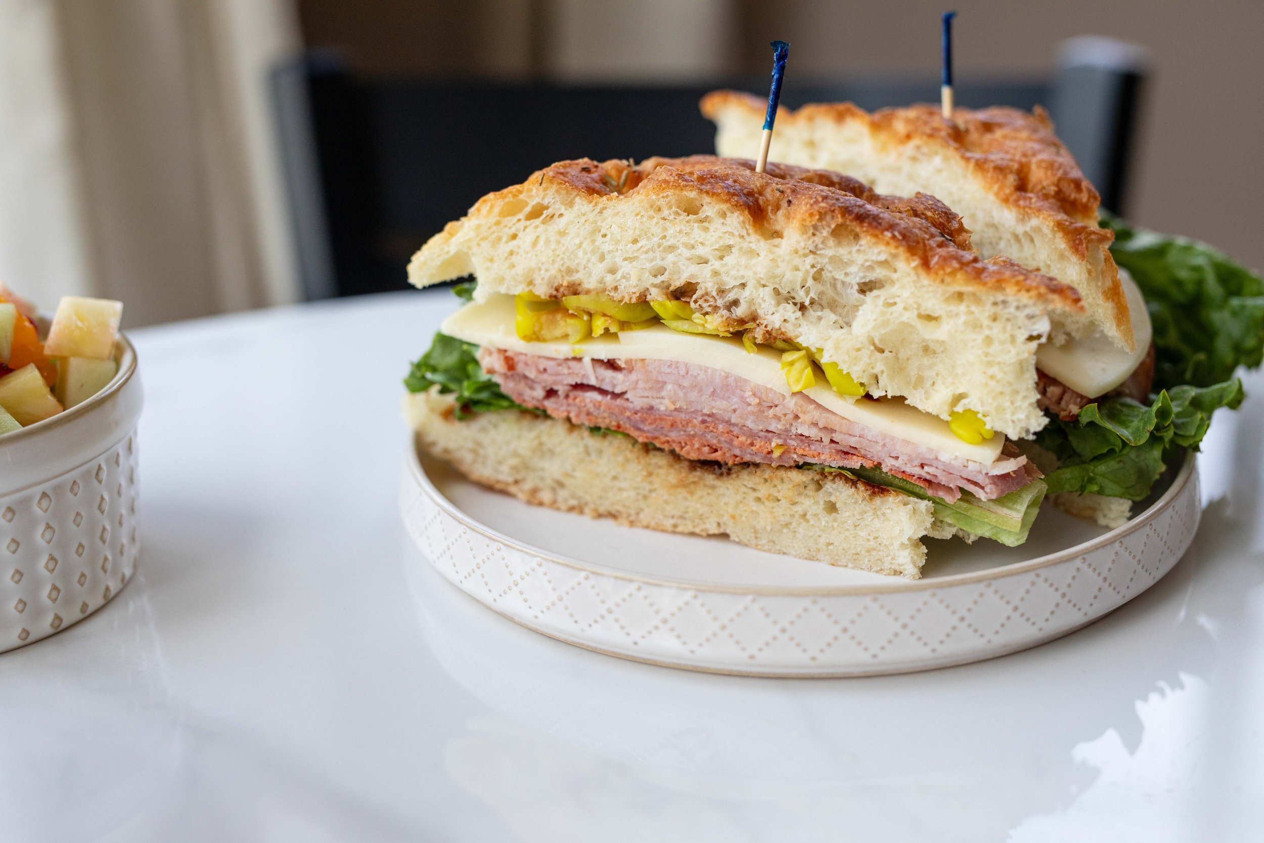 A sandwich with layers of deli meats, cheese, lettuce, pickles, and bread, held together with small decorative skewers, served on a white paper tray.