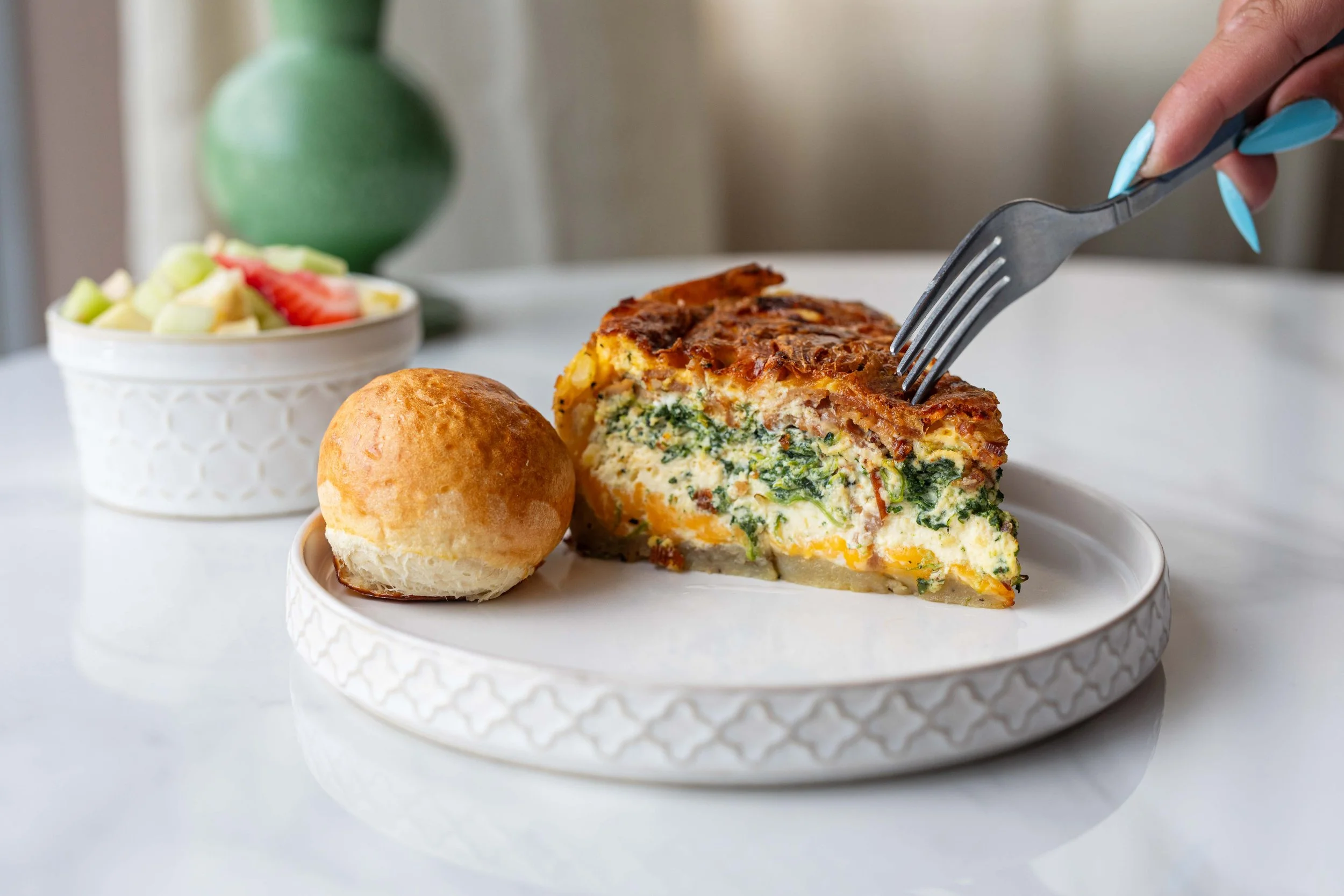 A slice of vegetable frittata on a white textured plate, garnished with a piece of toasted bread, with a bowl of potato salad and a green vase in the background.
