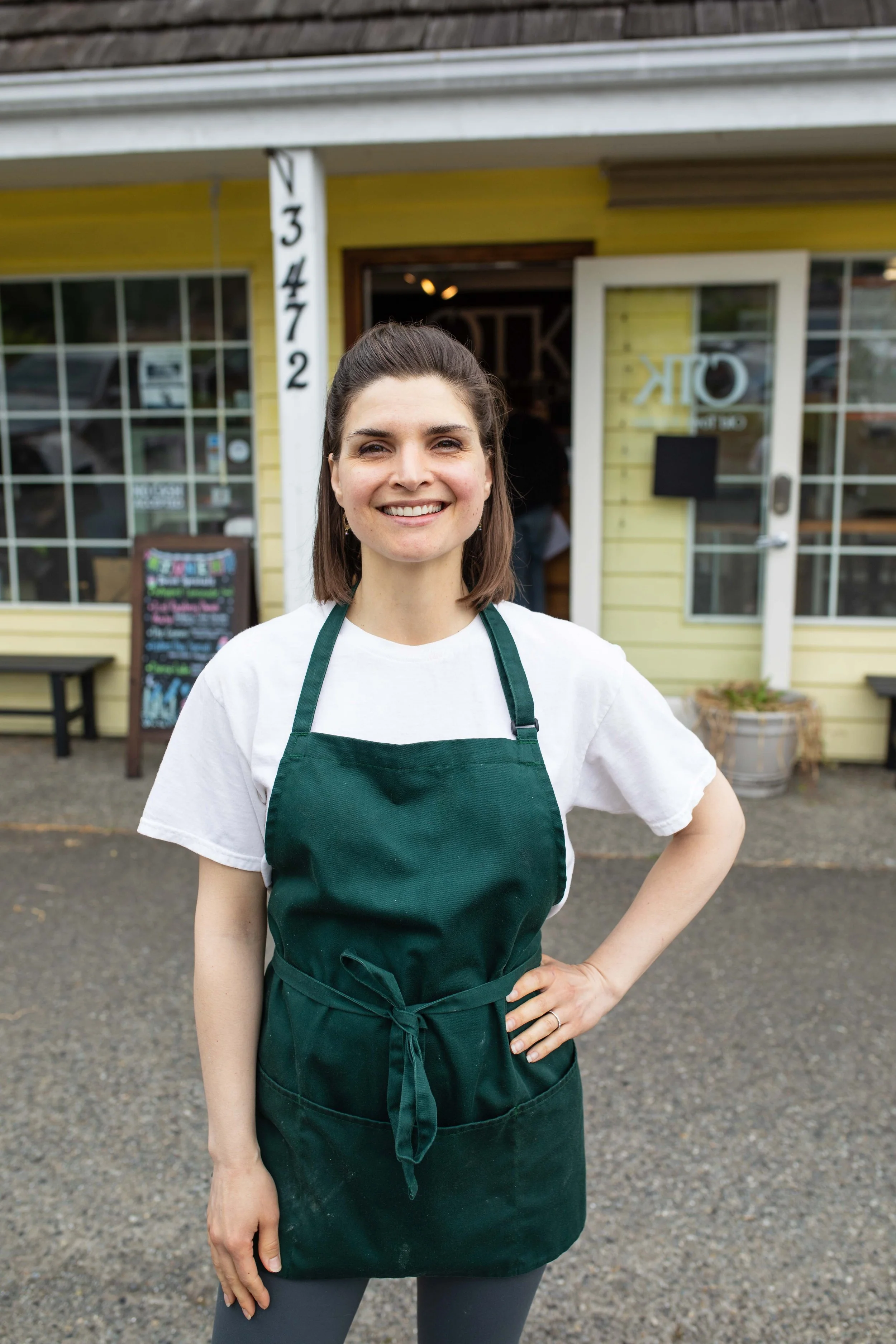 A smiling woman wearing a white t-shirt and a dark green apron standing outside in front of a yellow building with large windows and a door.