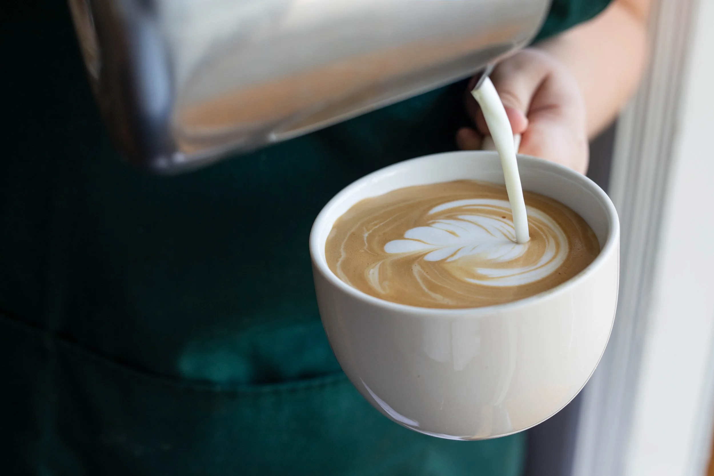 Close-up of a latte with foam art in a white ceramic mug on a dark green surface, with milk being poured from a silver pitcher.