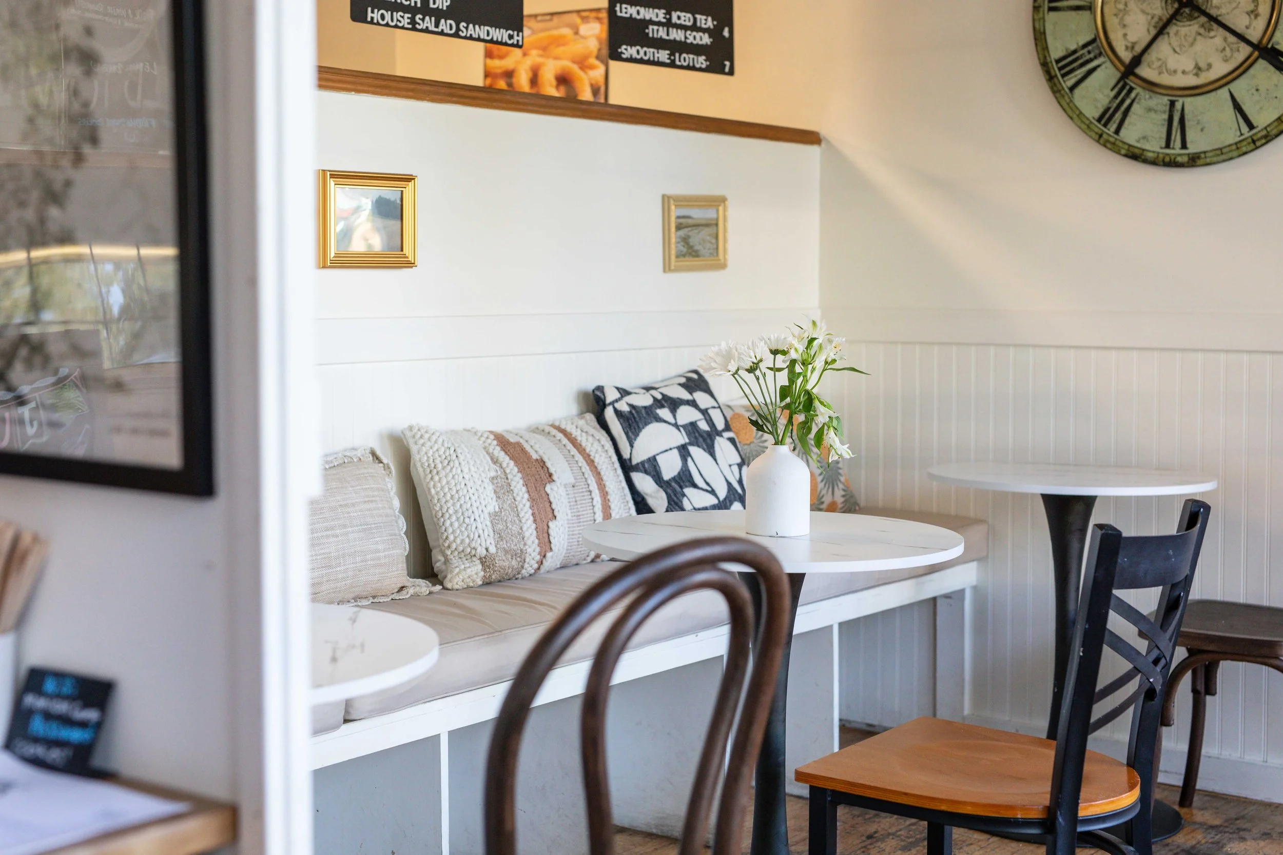 Interior of cozy cafe with a white bench seat decorated with patterned cushions, a round white table with a white vase of white flowers, surrounded by chairs, and wall art including framed pictures and a large vintage-style clock. A black menu board listing food and drink options is visible above.