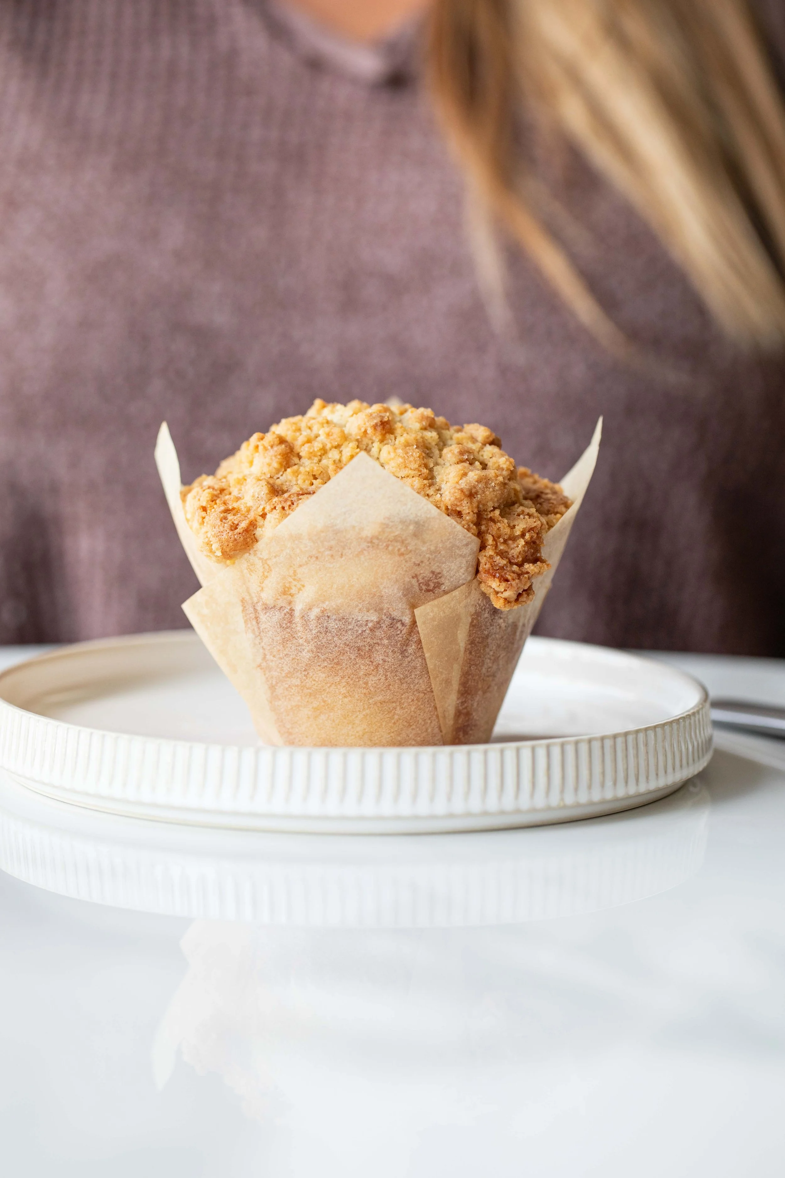 A close-up of a crumbly muffin in a paper wrapper, placed on a white textured plate against a blurred background of a person wearing a purple sweater.