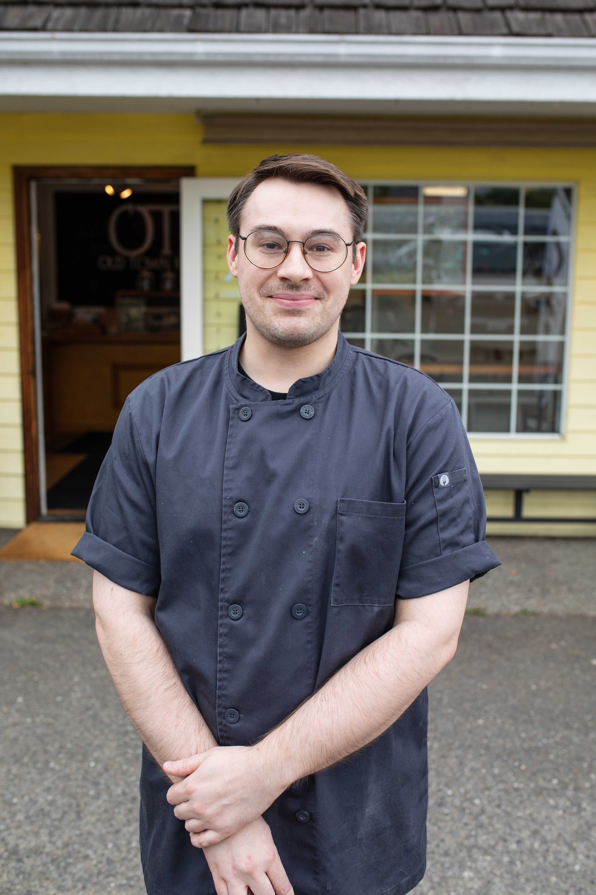 A young man with dark hair, glasses, and a slight smile, standing outside in front of a yellow house with a large window, wearing a dark gray chef's coat.