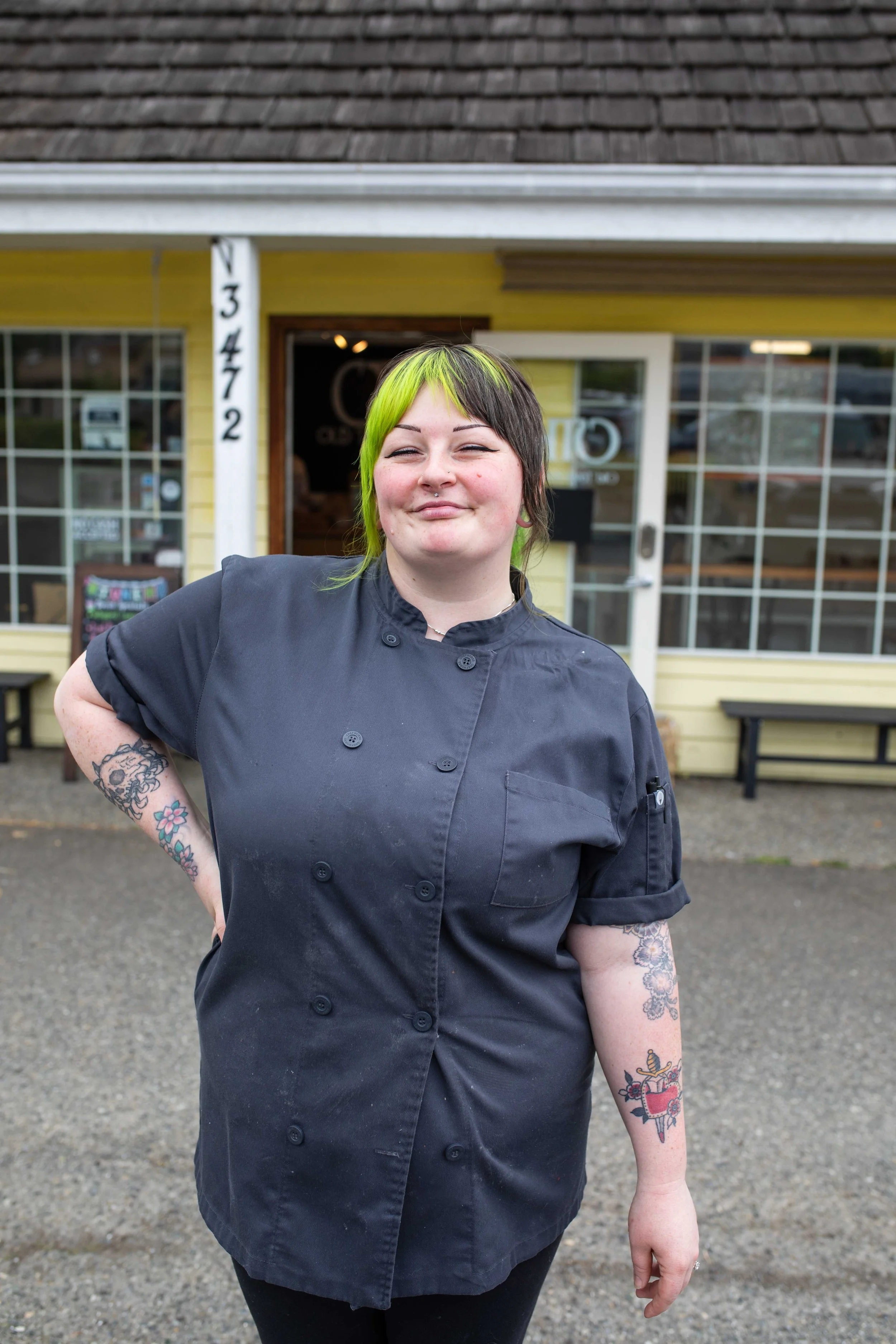 A woman with short dark hair with bright green front highlights, wearing a black chef's coat, standing outdoors in front of a yellow building with large windows and a gray shingled roof.