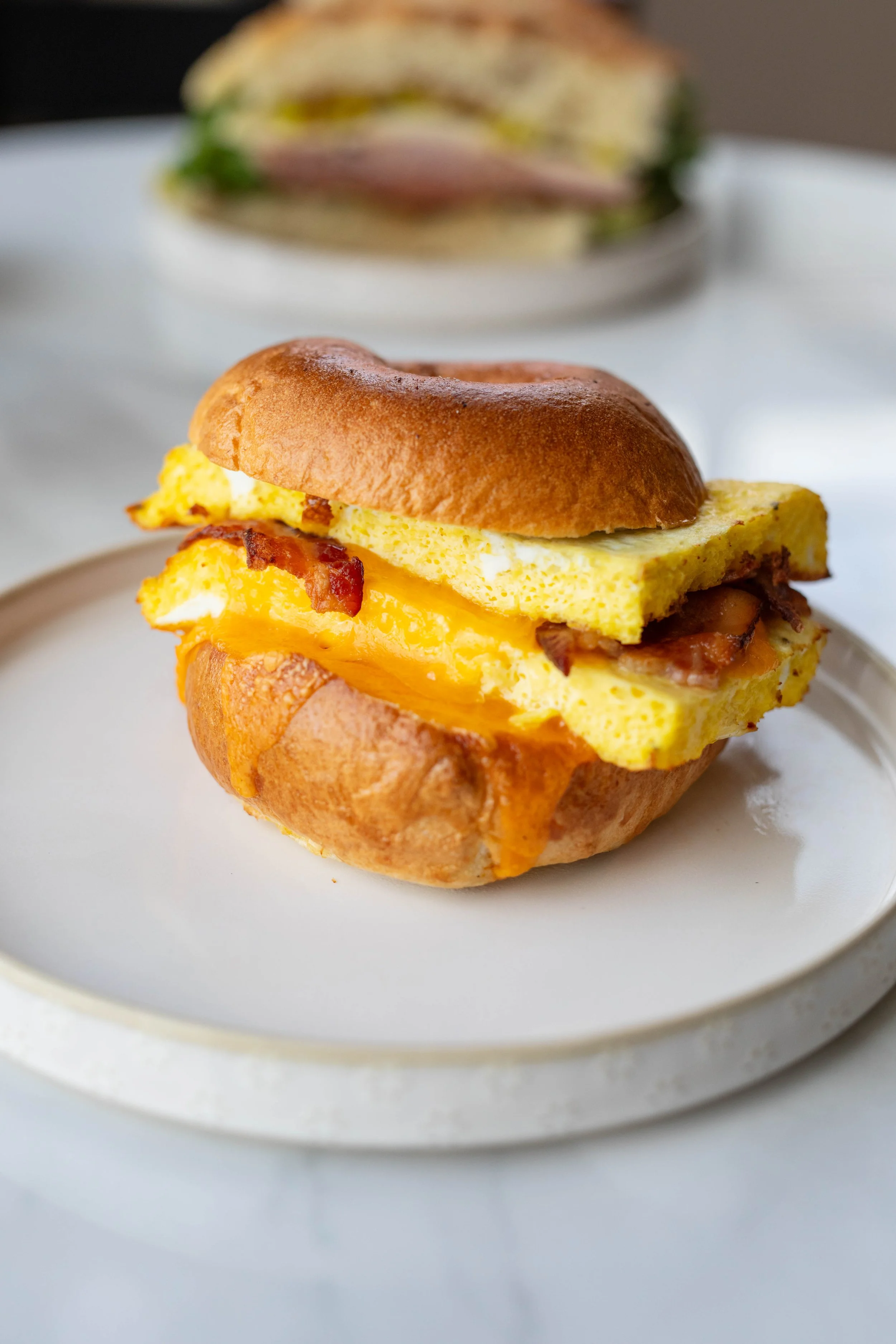 Close-up of a breakfast sandwich with scrambled eggs, cheese, bacon, and sandwich bun on a white plate.