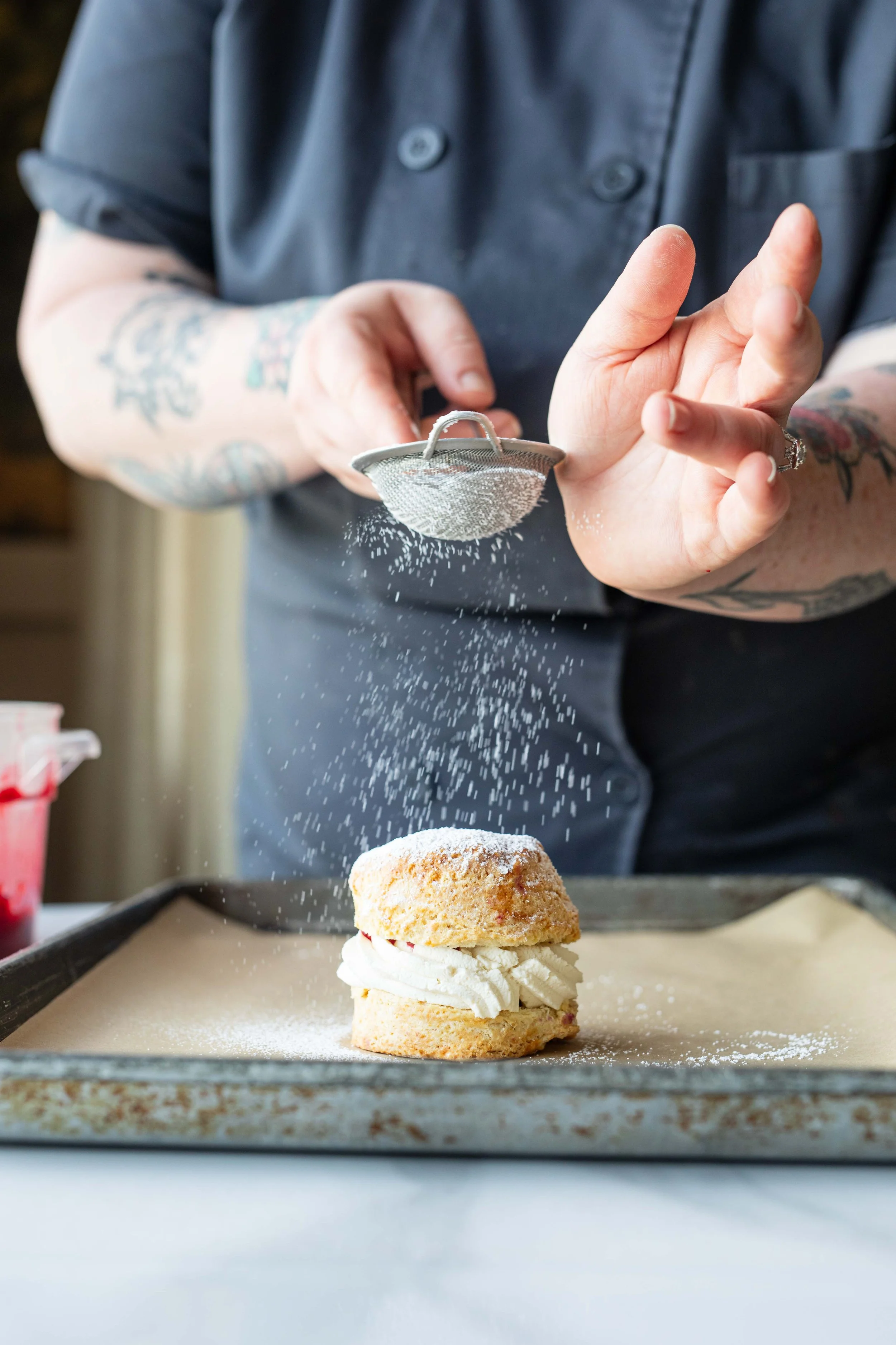 A person with tattoos on their arm sprinkles powdered sugar onto a dessert with cream filling, a biscuit, and powdered sugar on a baking tray.