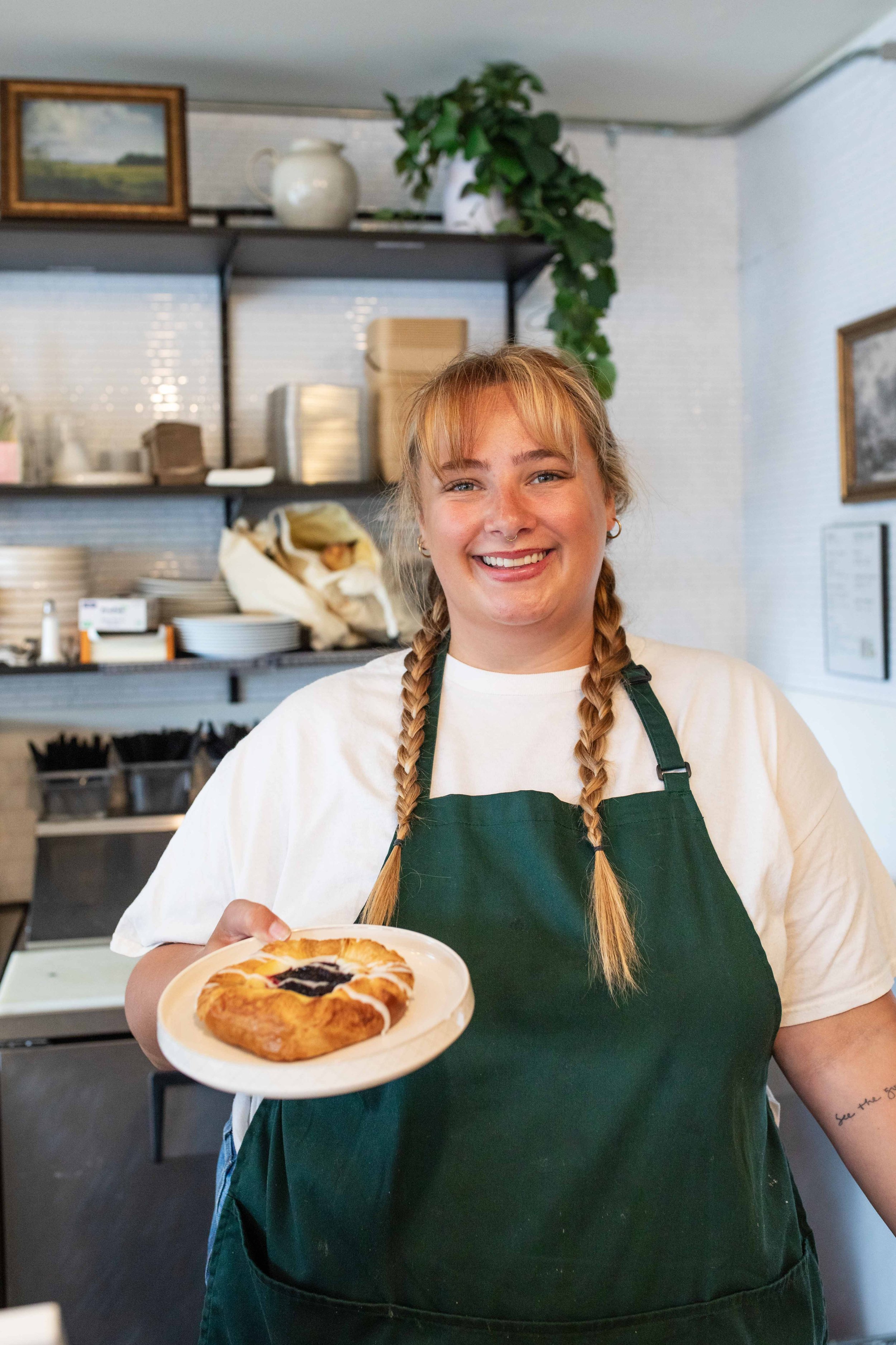 A young woman with blonde hair in braids, wearing a white t-shirt and green apron, holding a plate of baked good, smiling in a cafe setting.