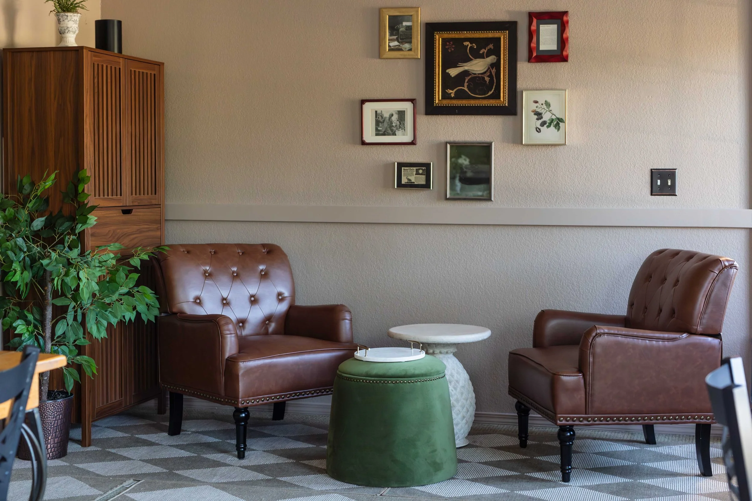 A cozy sitting area with two brown leather armchairs, a small round marble-top table, and a green upholstered stool. There is a gallery wall with framed artwork and photos on the beige wall. A wooden cabinet with plants on top is visible on the left, and the floor has a patterned carpet.