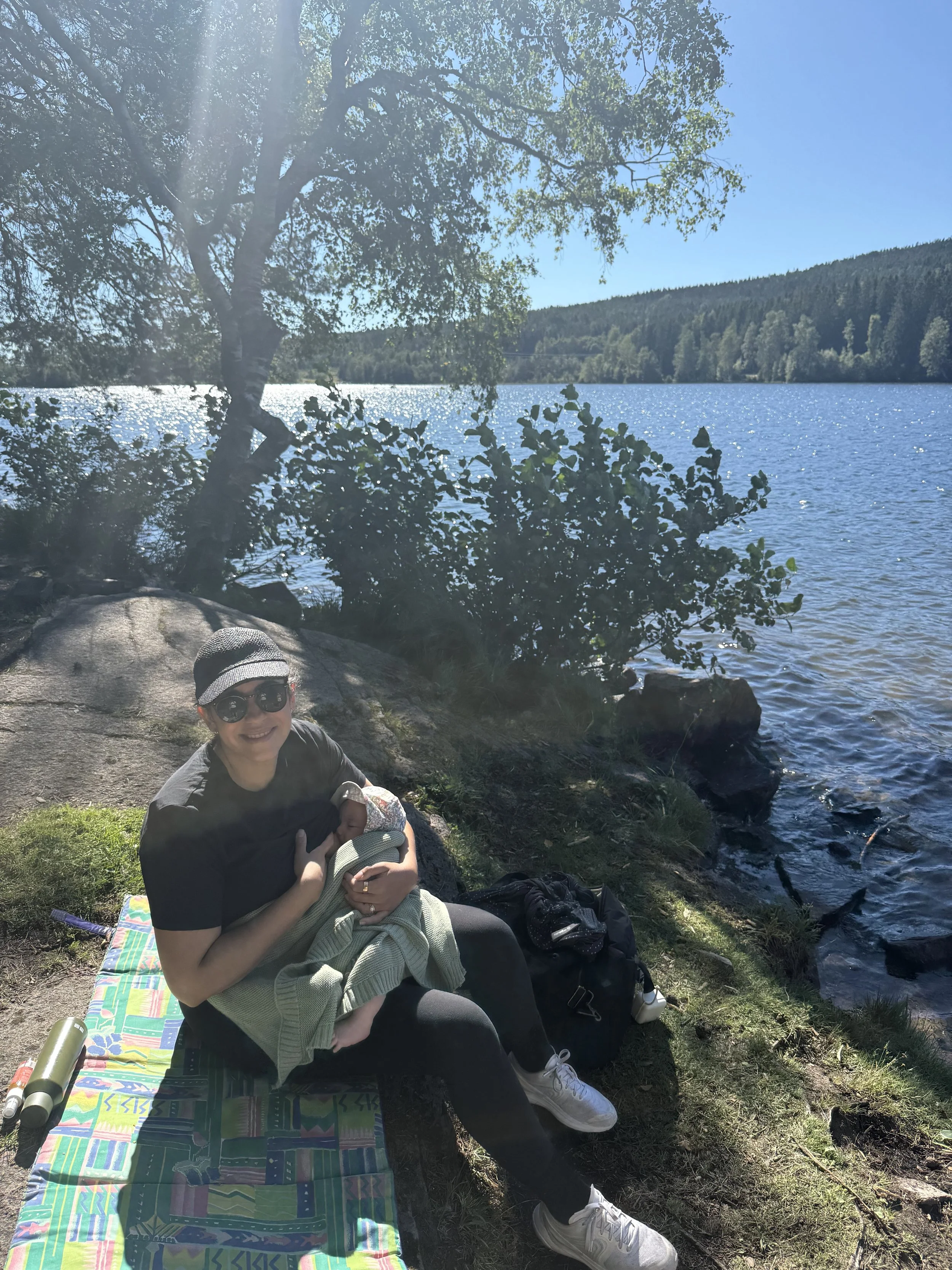 Woman sitting on a colorful blanket by a lake, holding a baby, with trees and water in the background on a sunny day.