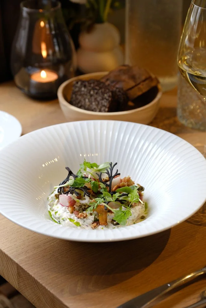 A white bowl containing a finely garnished dish with herbs, microgreens, diced ingredients, and a creamy base, placed on a wooden table. In the background, there are slices of bread in a bowl, a candle, a candle holder, a glass of white wine, and a glass of beer.