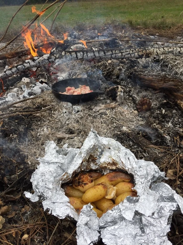 Een kampvuur met een gietijzeren pan met vlees erboven en een stuk aluminium folie met geroosterde uien.