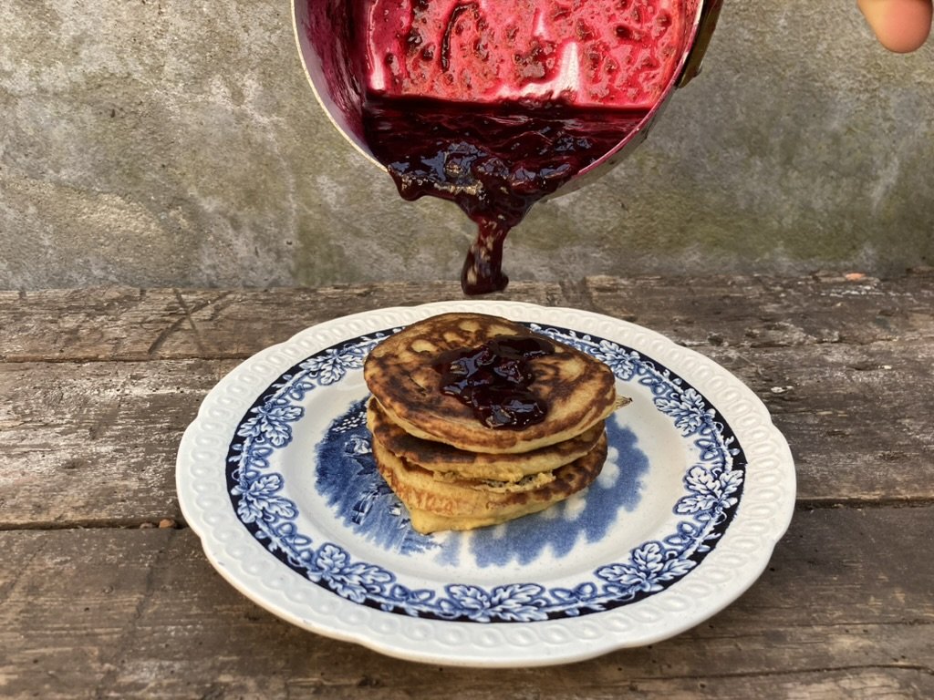 een stapel van drie gouden gebakken pannenkoeken op een blauw en wit bord, met bosbessenjam eroverheen en een blauwe saus ernaast, alles op een houten tafel.