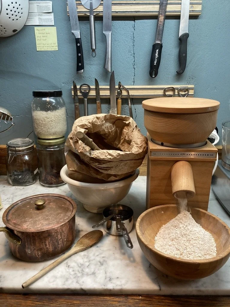 Werkbank met keukengerei en ingrediënten, waaronder een molen die gemalen graan aan een kom toevoegt, een houten pan, een glazen pot met rijst, en verschillende messen en schaartjes aan de muur.