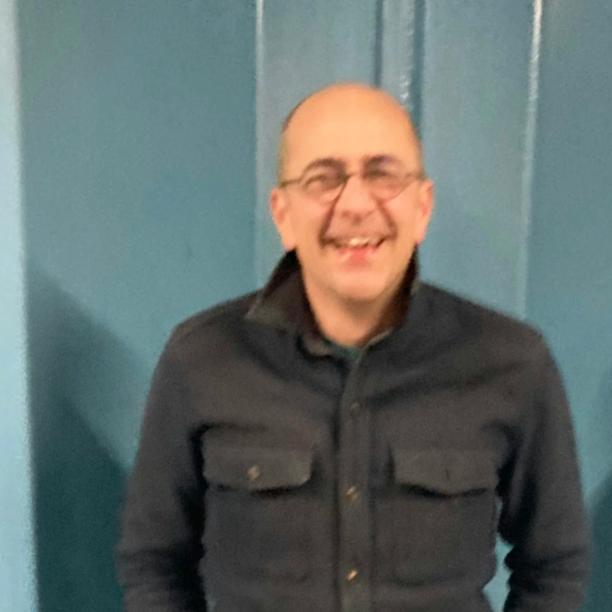 Man met zwarte haar en een baard, draagt ronde glazen en een zwarte trui, zit in een lichte kamer met een plant op de achtergrond.