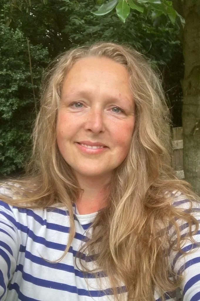 Een vrouw met lang, golvend blond haar, die een glimlach op haar gezicht heeft, buiten met groene bladeren op de achtergrond.