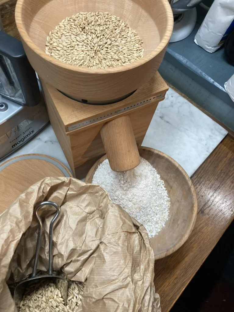 Op een tafel staan een houten molen met graankorrels, een grote houten kom met gemalen graan en een papieren zak met nog meer meel en een metalen lepel.