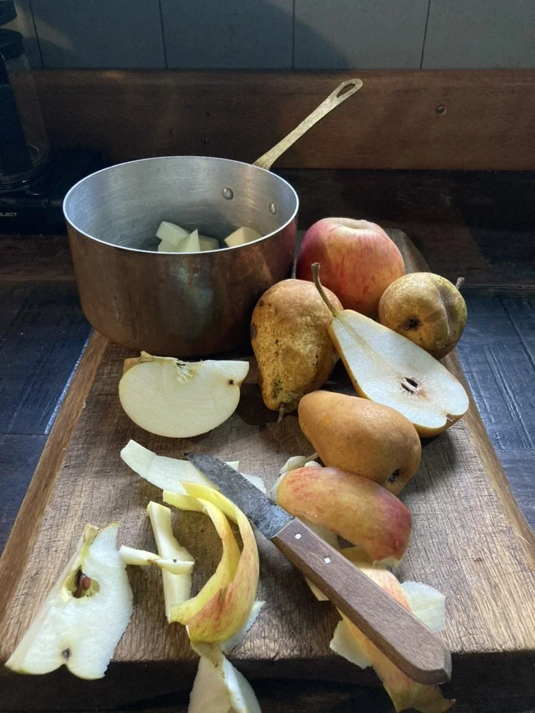 Een houten snijblok met verschillende neuspeertjes en een mes, een pan met gesneden aardappels, en verschillende gesneden en hele peren en appels.