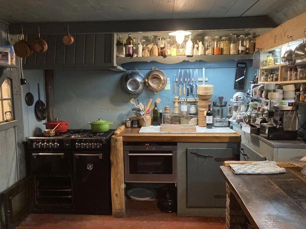 Een knusse keuken met een kookplaat, groene en rode potten, open planken met potjes, messen en keukengerei, een snijblok, oven, en verschillende keukentoestellen en container voor kruiden.