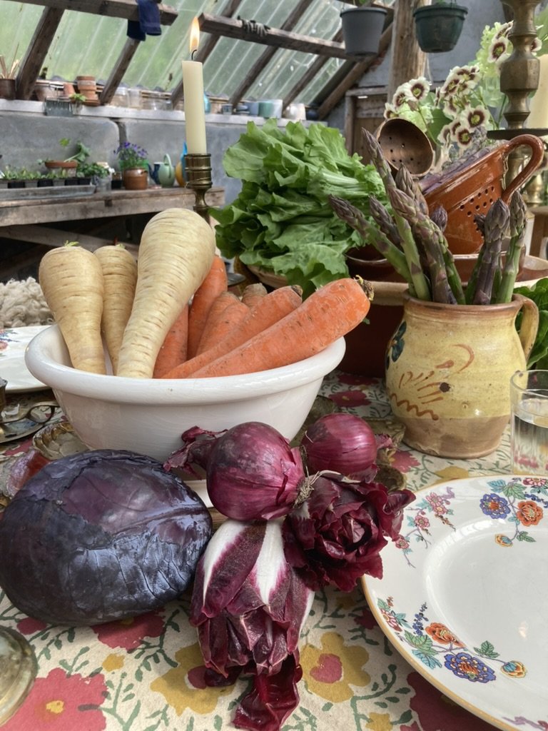Een tafel met verse groenten zoals pastinaak, wortel, rode ui, rode kool, en artisjok, alsook een vaas met asperges en een ander met bloemen; er is ook een kaars en een bord met bloemenpatroon.