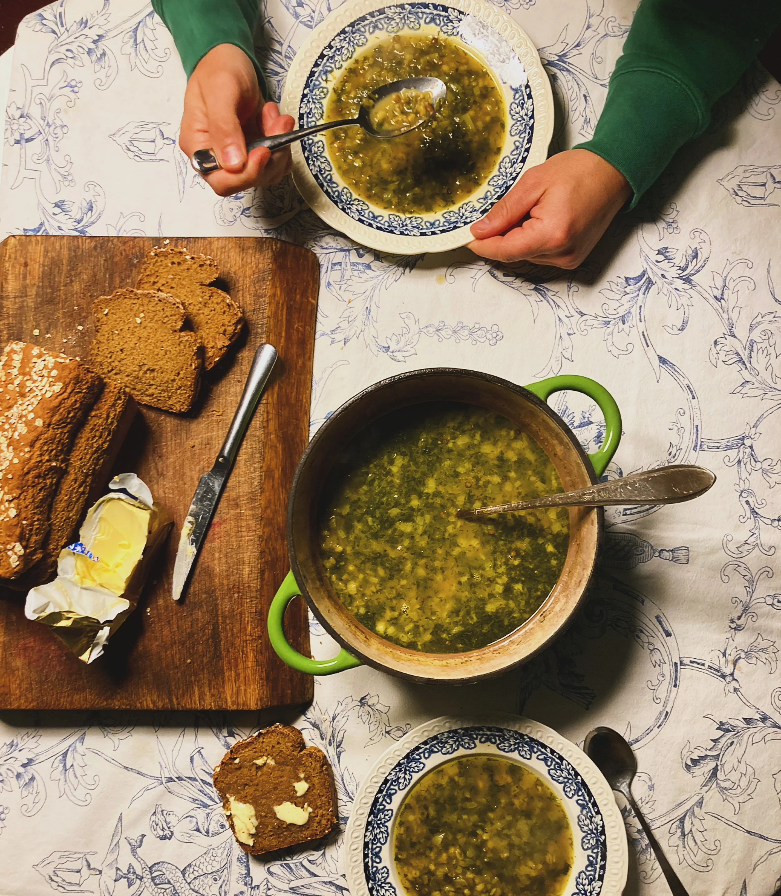 Een tafel met twee kommen hete groene soep, een houten snijplank met volkorenbrood, een stuk boter en een mes.