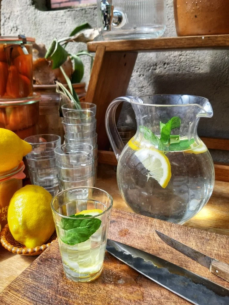 Een glas water met citroen en munt, een fles met water, drie lege glazen, en twee citroenen op een houten snijplank.