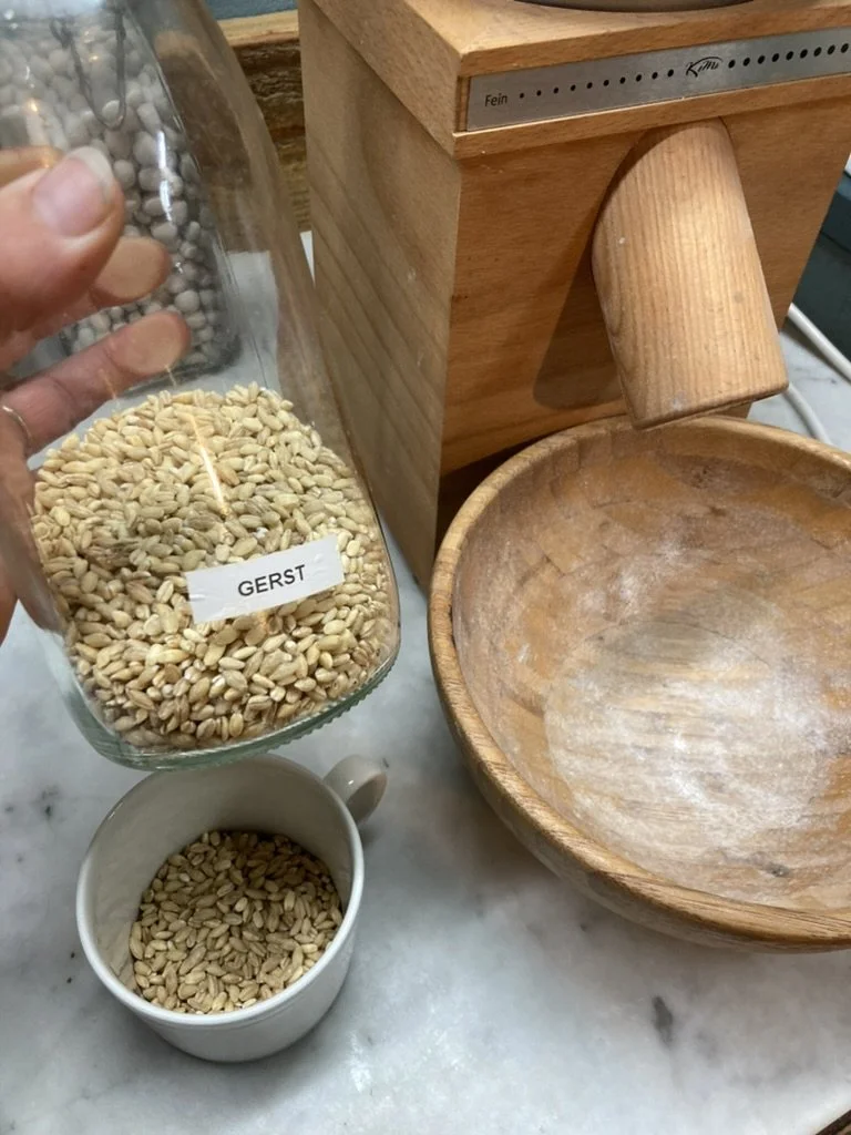 Een hand die gerst (Gerst) uit een glazen pot schenkt in een witte kop, op een keukentafel naast een houten molen en een lege houten kom.