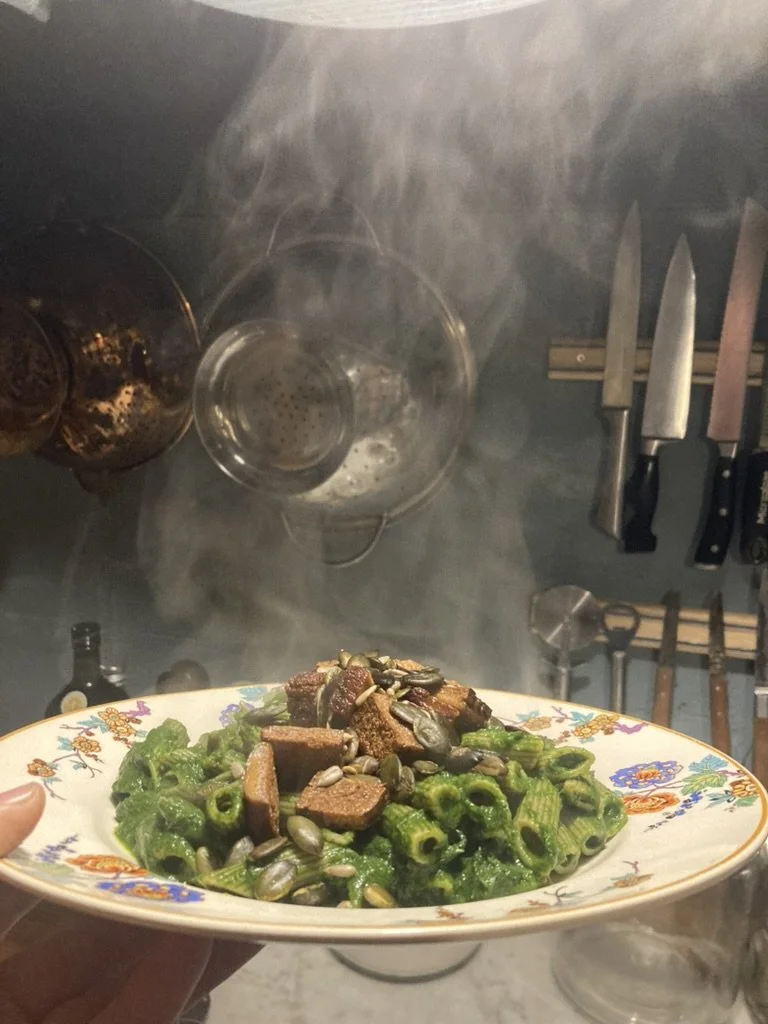 Een bord met groene pasta, stukjes vlees, en zaden, vastgehouden door iemand. In de achtergrond bevinden zich keukengerei en glazen, met stoom die opstijgt.