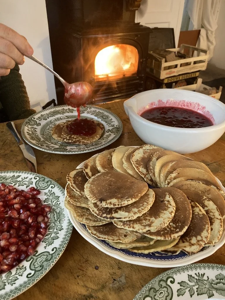 Een tafel met pannenkoeken, een kom met bessensaus, een bord met granaten, een haard met open vuur, en iemand die aardbeienjam op een pannenkoek schept.