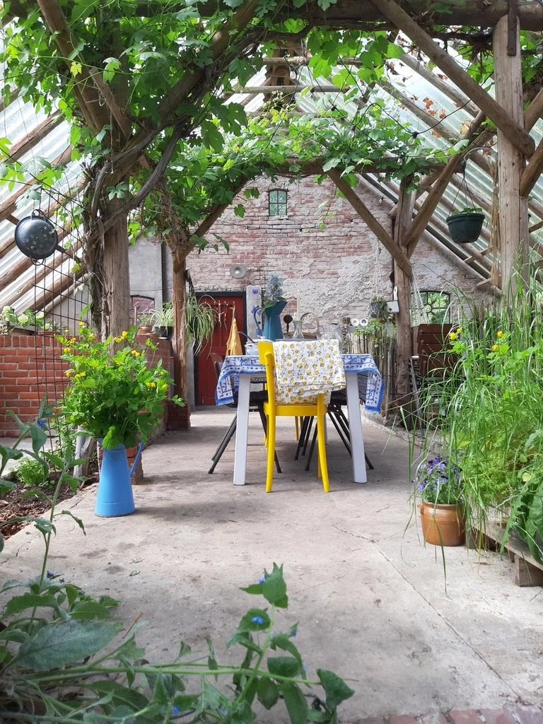 Een gezellige tuin met een eettafel, rondom gekleurde stoelen, onder een boom met groene bladeren in een kas-achtige structuur met glas en hout. Er zijn diverse planten en bloemen in potten en vazen.