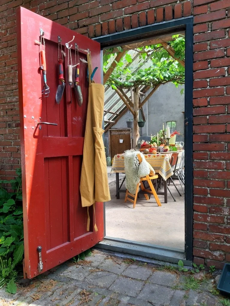 Open rode tuinpoort die uitkomt in een buitenruimte met een eettafel, versierd met fruit, kaarsen en servies, onder een houten pergola met groene bladeren, en omgeven door bakstenen muren.