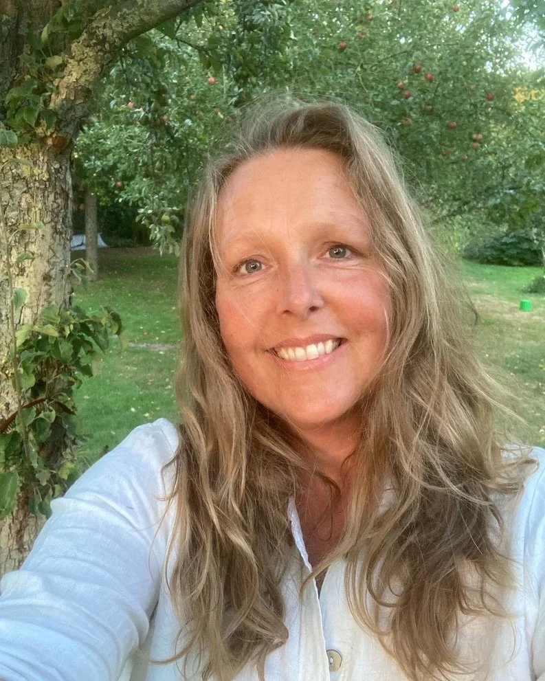 Een vrouw met lang, golvend haar die buiten voor een boom met fruit poseert, lachend naar de camera.