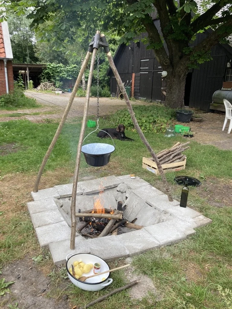 Een open haard met een pot op een hengel erboven en een vuur in de haard. Naast de haard ligt een kom met aardappels en een houten lepel. Op de achtergrond ligt een hond op het gras onder een boom, met wat speelgoed en een houten krat in de tuin.