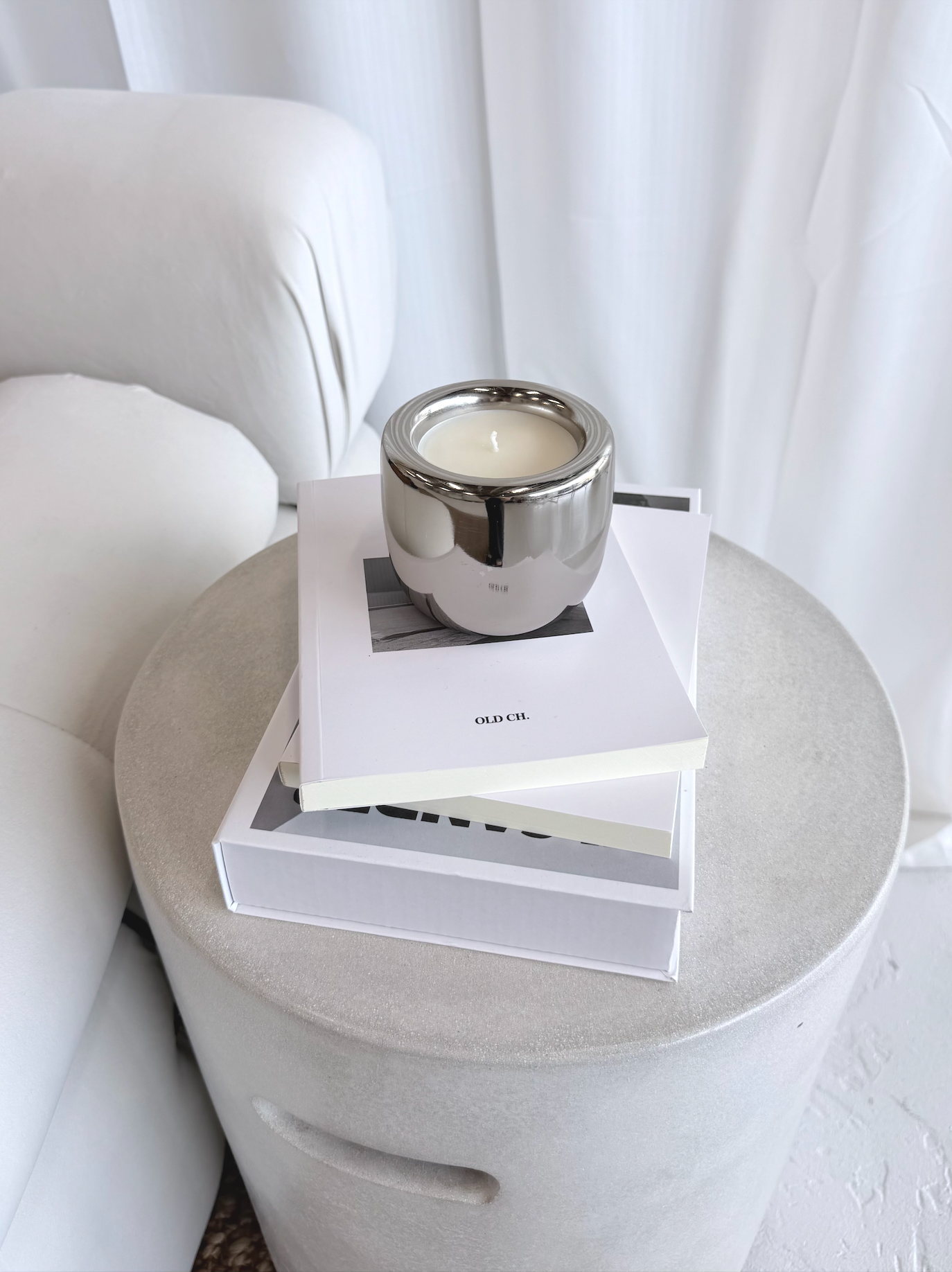 A silver candle holder with a lit white candle on top of two stacked white books, placed on a white table with a light-colored, rounded surface, against a background of white curtains.