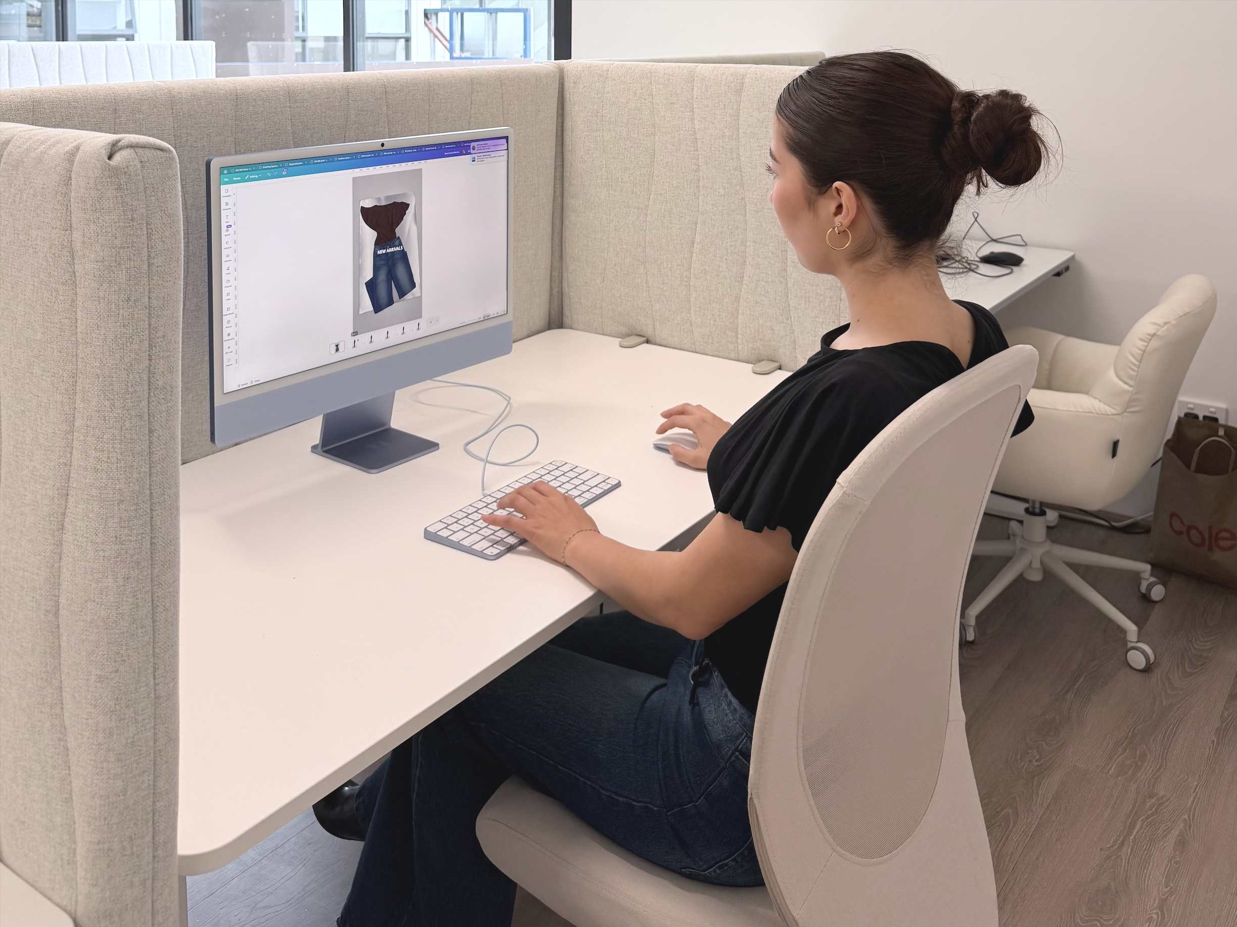 A woman sitting at a desk in an office, working on a computer with a design of blue jeans on the screen, wearing a black top and earrings, with a partition and other office furniture in the background.