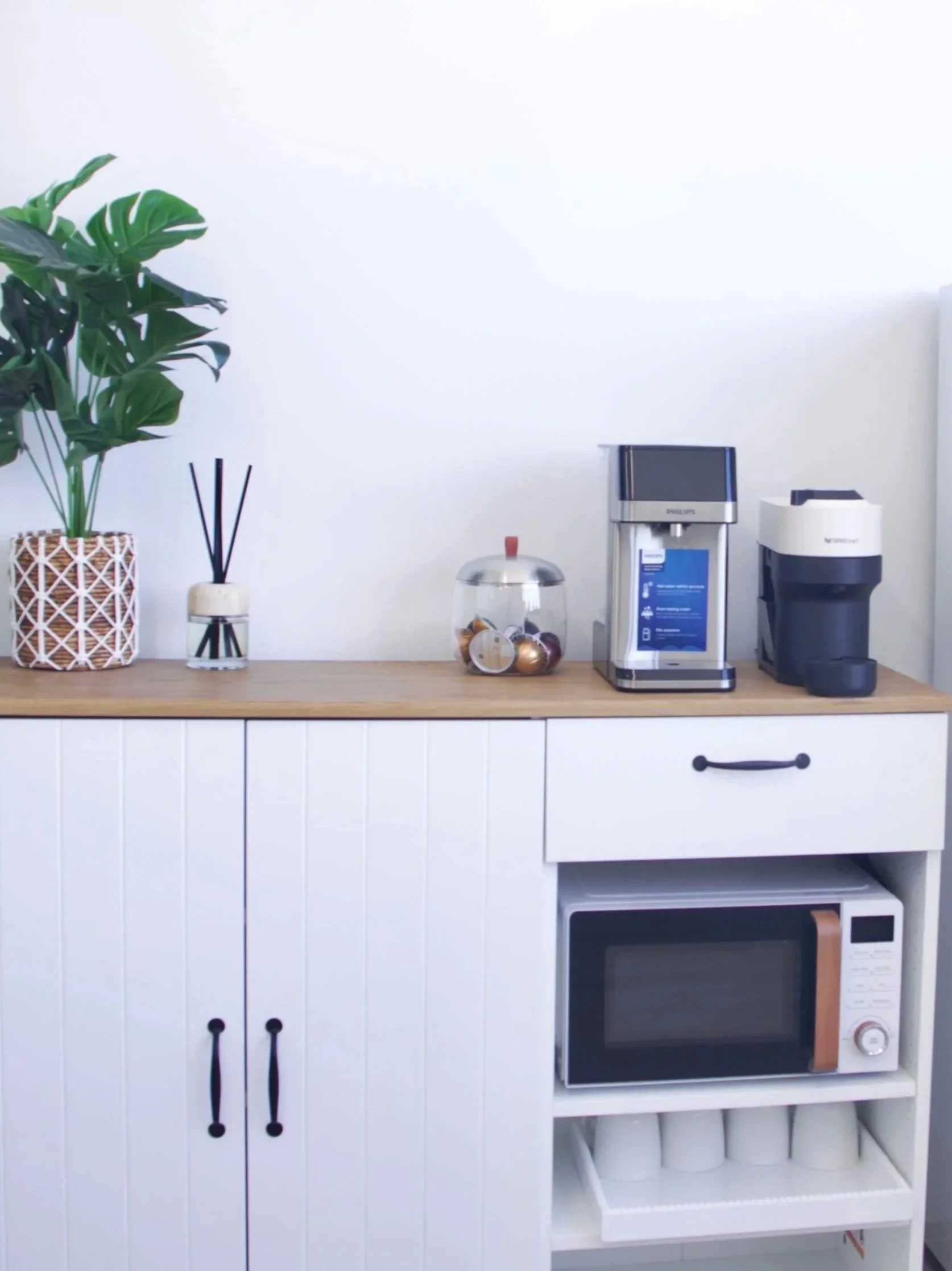 Kitchen countertop with a potted plant, a reed diffuser, a jar with candies, a water dispenser, a coffee machine, a microwave oven, and a paper towel roll