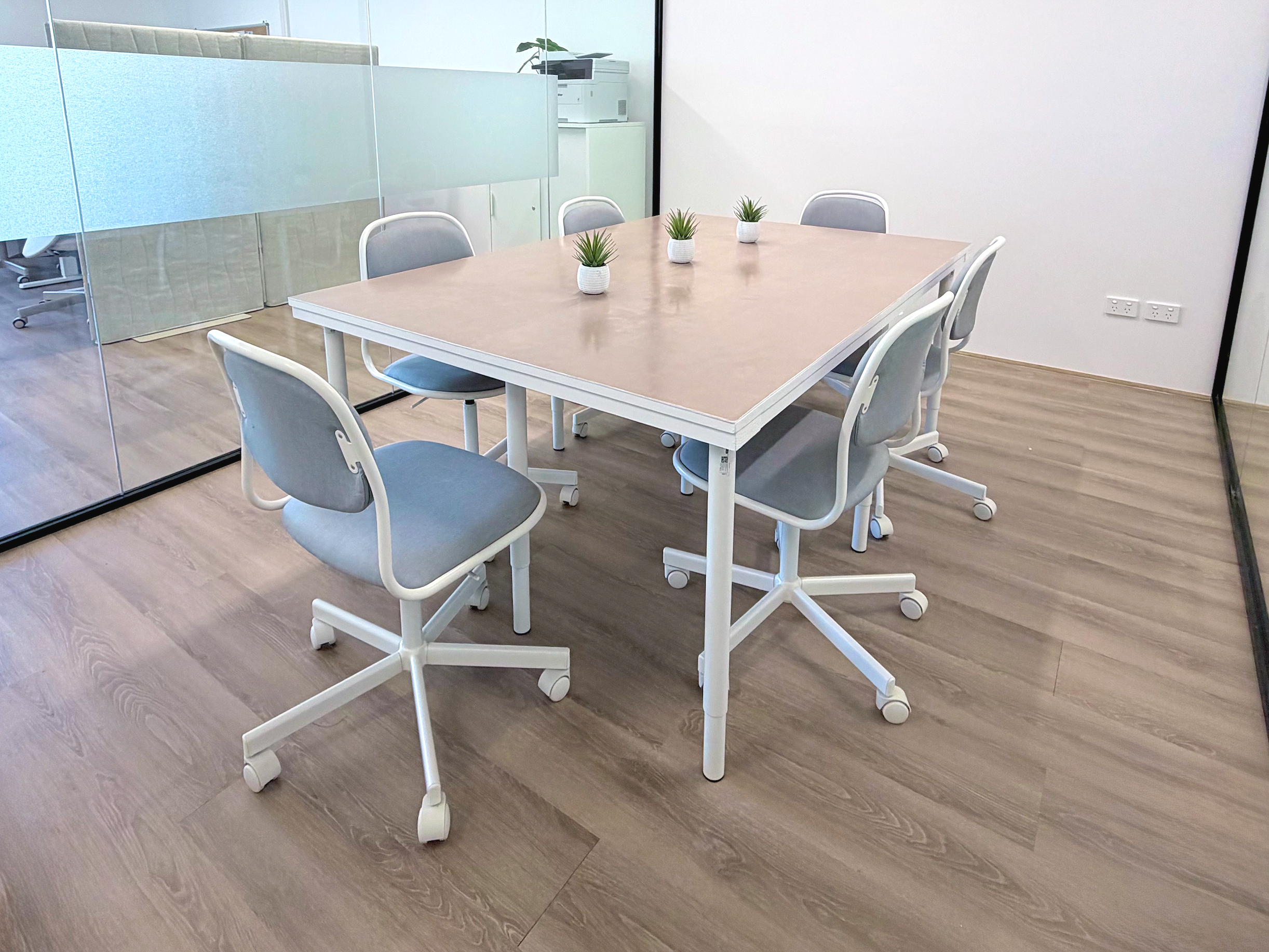 A conference room with a wooden table, six chairs with gray upholstery, and three small potted plants on the table. The room has a glass partition on one side and a white wall with electrical outlets on the other.