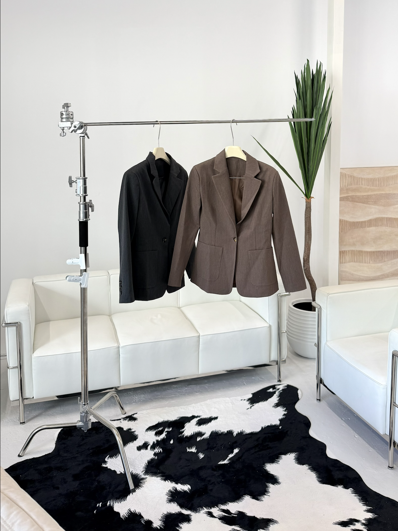 Two blazers, one black and one brown, hanging on a clothing rack in a modern interior with a white sofa, a cowhide rug, and a large green plant in a white pot.