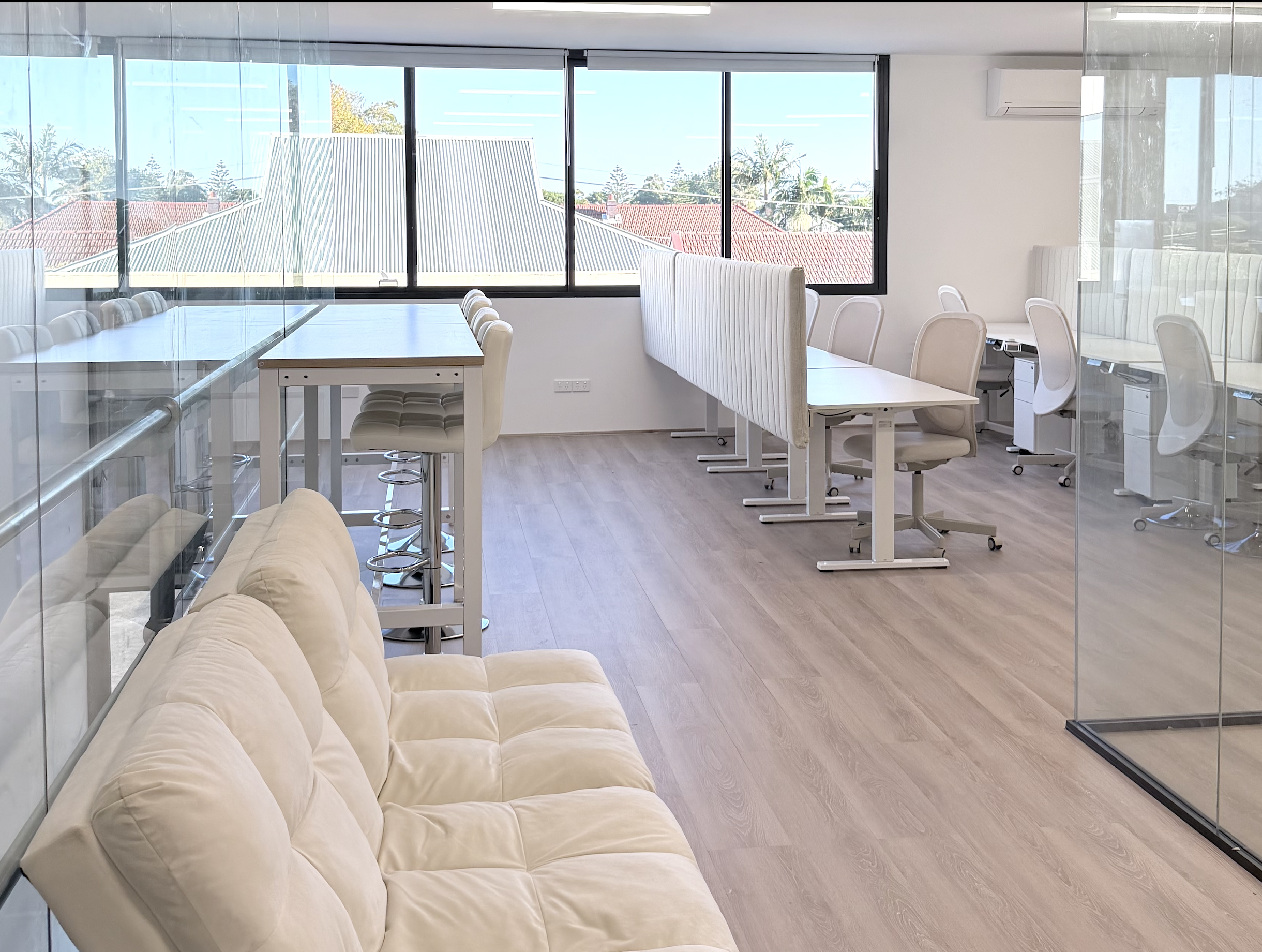 An office with large windows showing rooftops and trees outside. The office has a beige sofa, white cubicle desks with white chairs, and a wooden table near the window. There is an air conditioning unit on the wall.