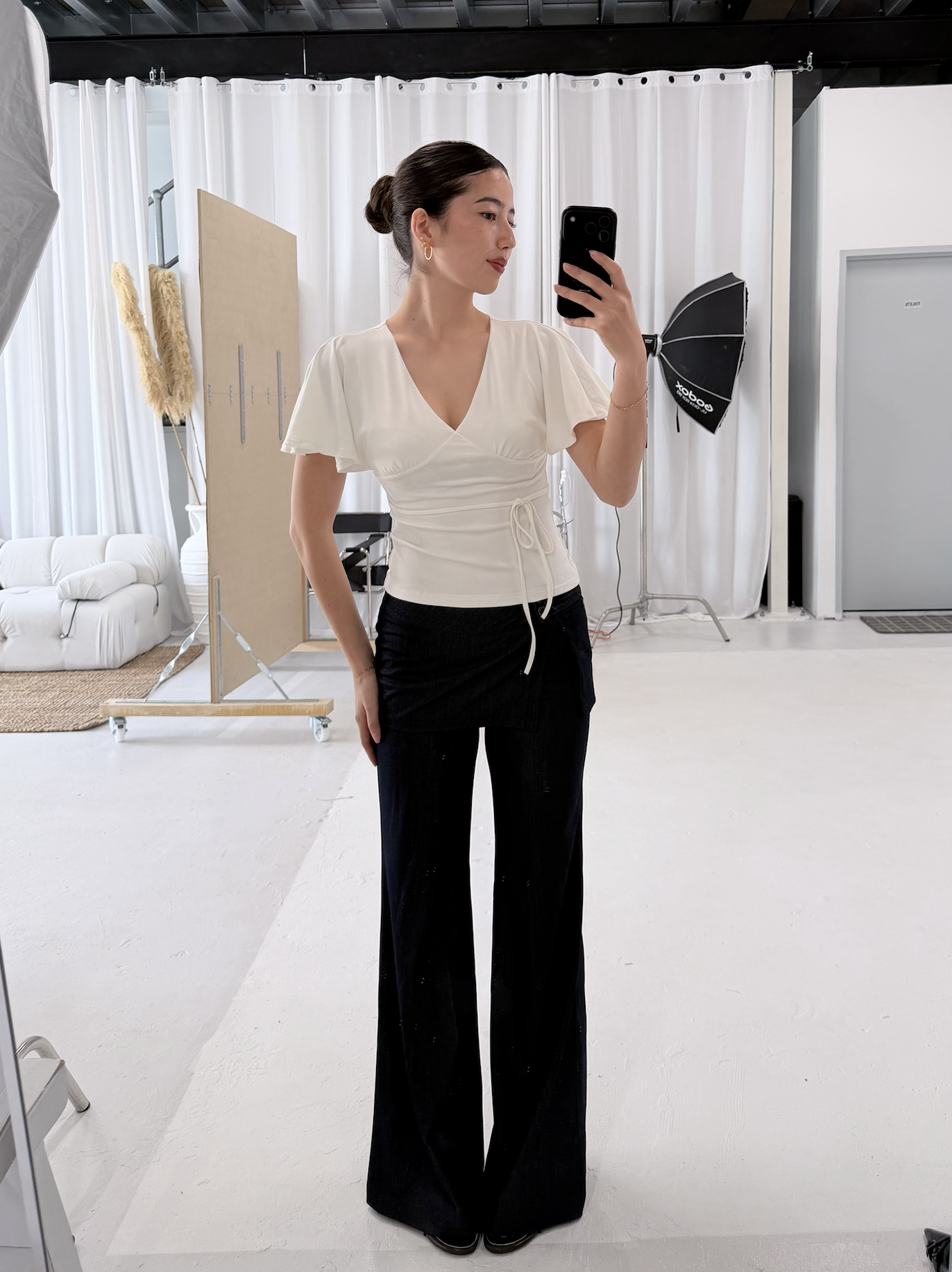 A woman taking a mirror selfie in a photography studio. She is wearing a white top and black pants, with her hair tied back.