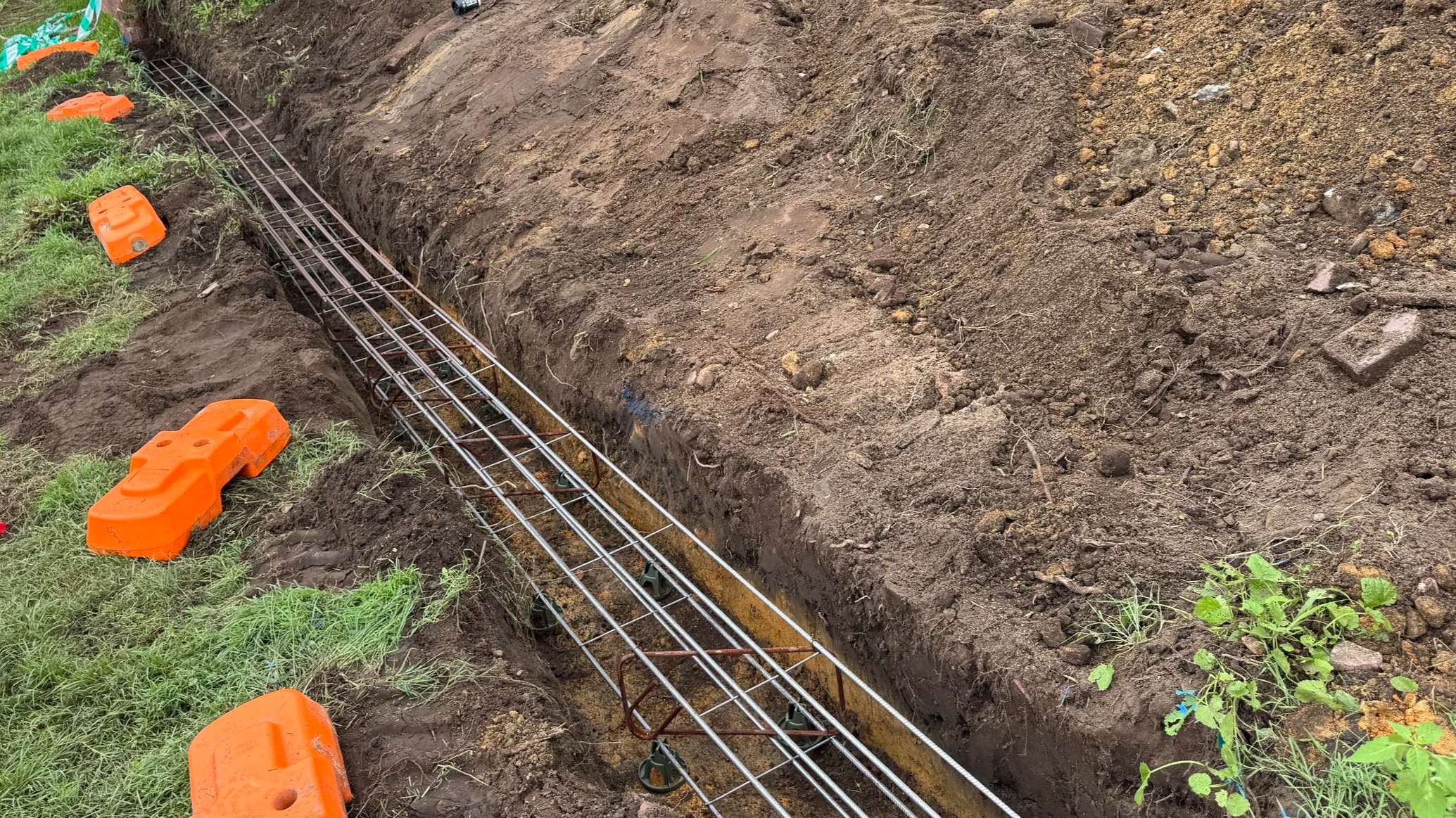 Construction site with metal rebar placed in a trench and orange safety barriers around the edges.
