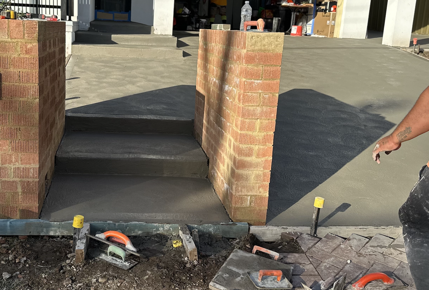 Construction site showing freshly poured concrete steps and sidewalk, brick walls, tools, and a worker's tattooed arm visible.