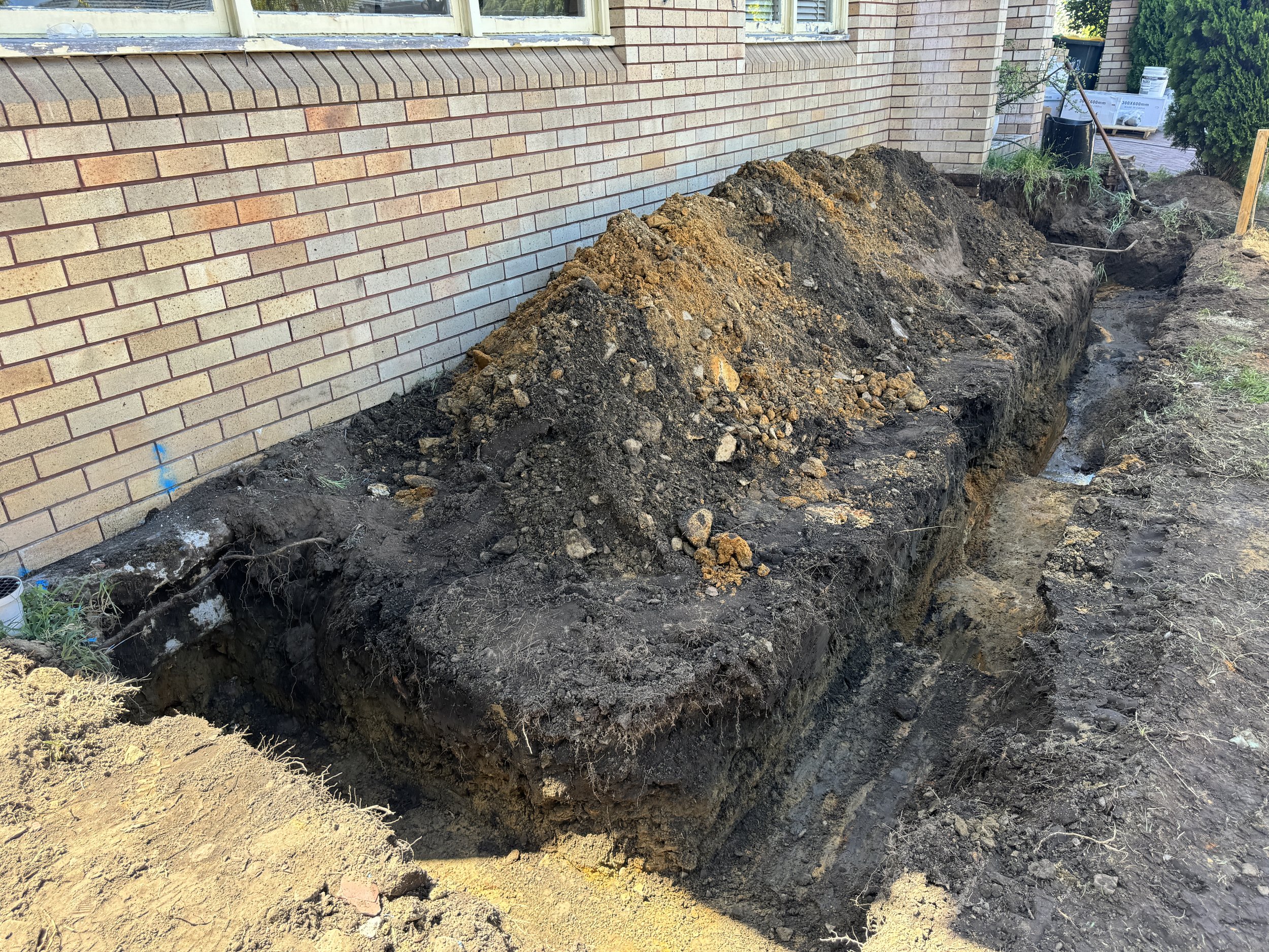 A construction site with excavated trench alongside a brick house wall, containing dirt, rocks, and construction tools.